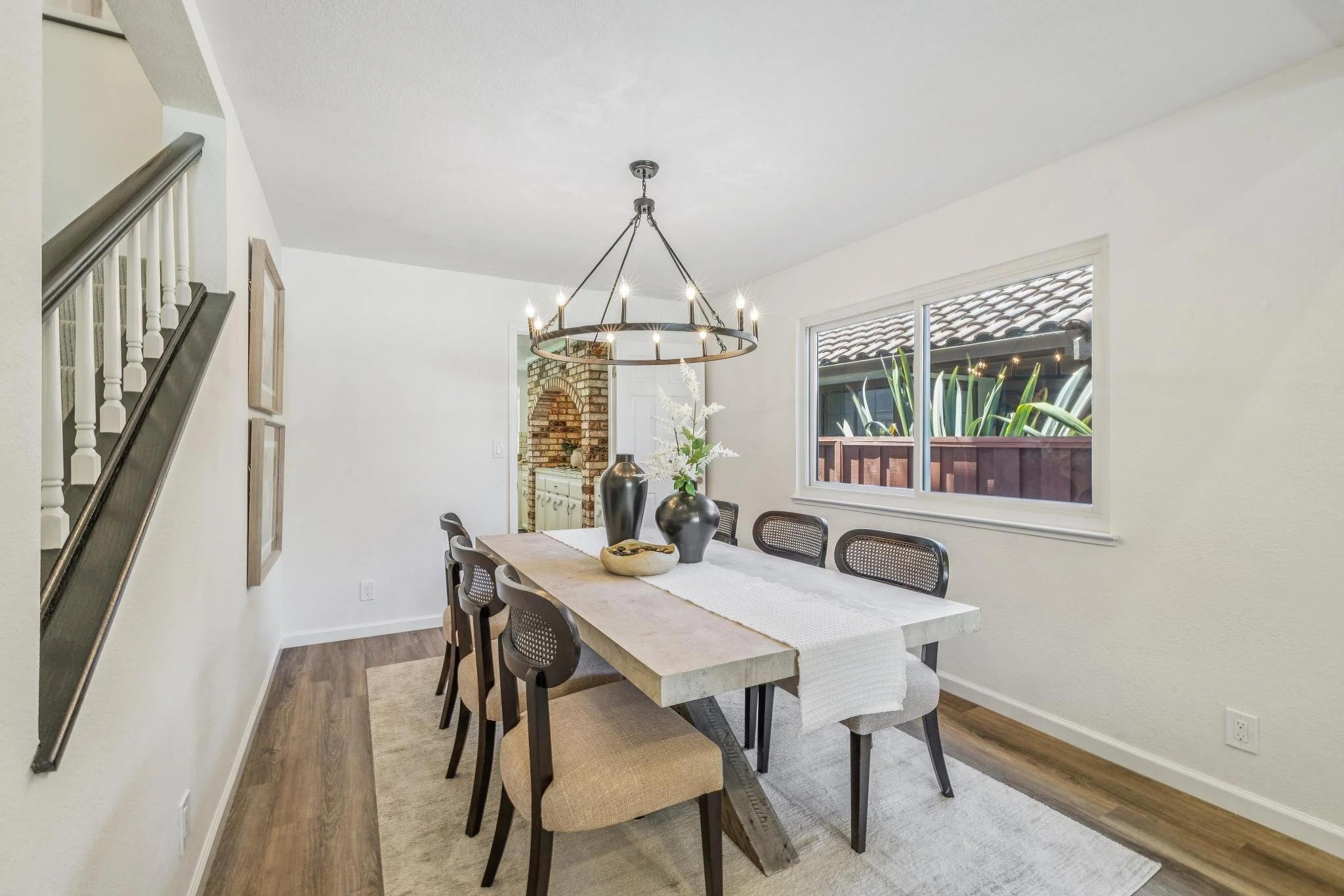 Dining room with a large wooden table, six chairs, a modern chandelier, floral centerpiece, window with an outdoor view, and a brick fireplace in the background.