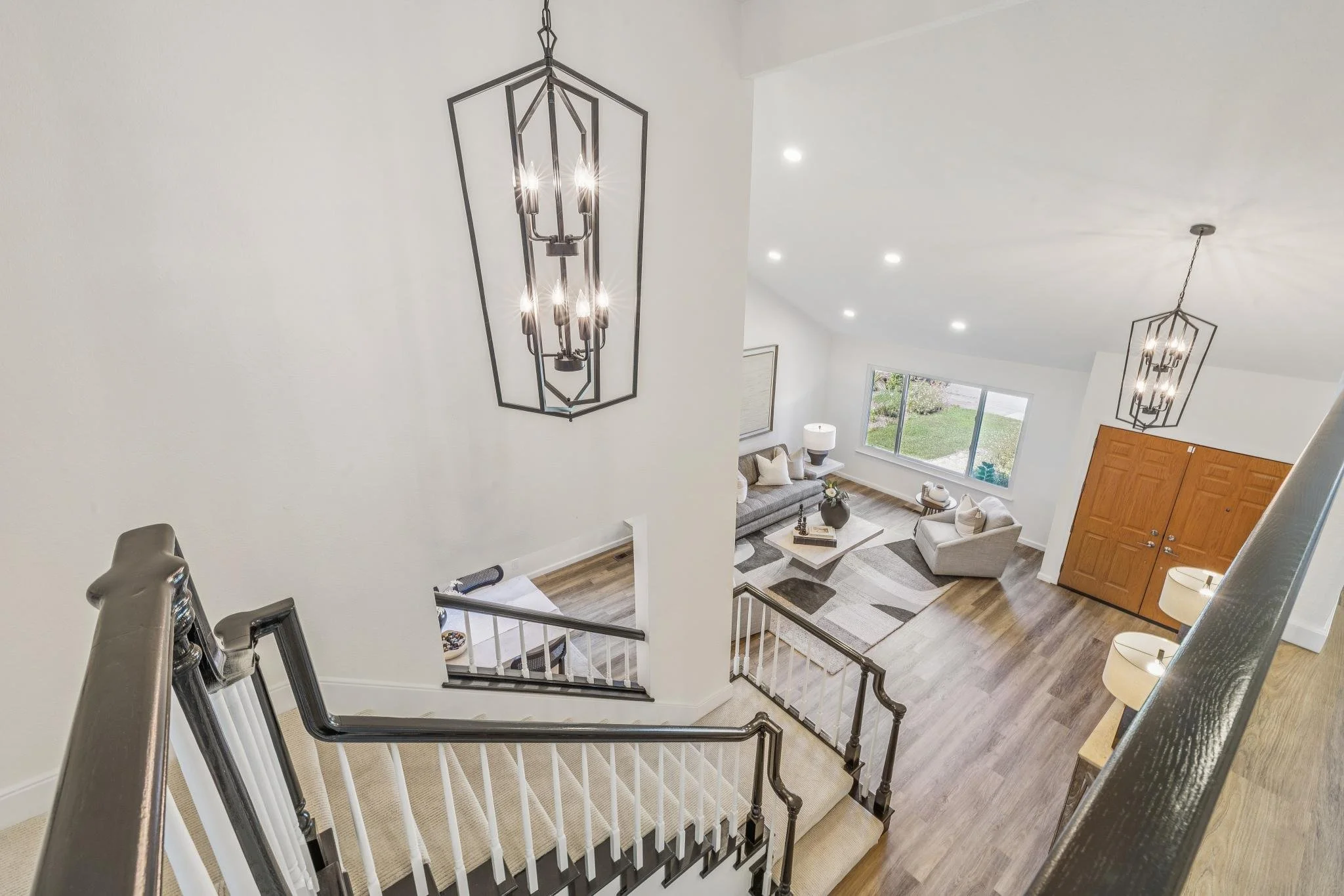 Interior view of a living room and staircase from above, with modern lighting fixtures, large window, white walls, and wood flooring.