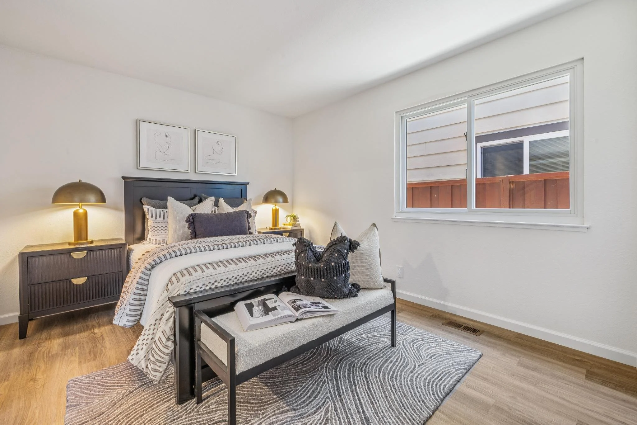A cozy bedroom with a bed, two nightstands with gold lamps, artwork above the bed, a small bench at the foot of the bed with an open book, a window with white blinds, and a patterned area rug.