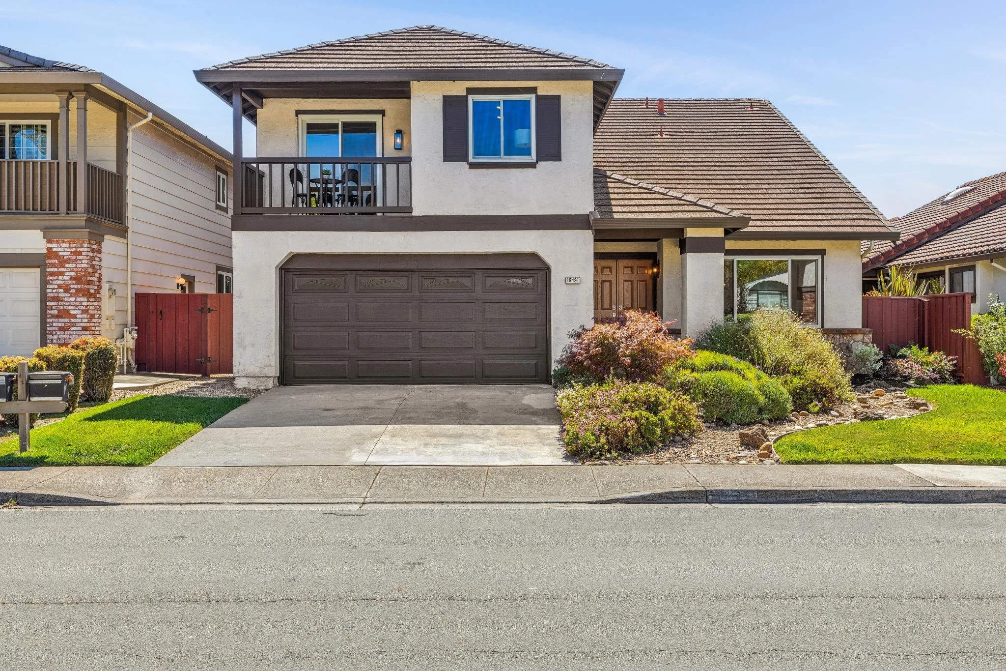 Front view of a two-story house with a gray garage door, a wooden front door, and a small balcony. The house has white walls and brown trim, with a sloped roof. There are landscaped bushes and flowers in front of the house, and the driveway and sidew