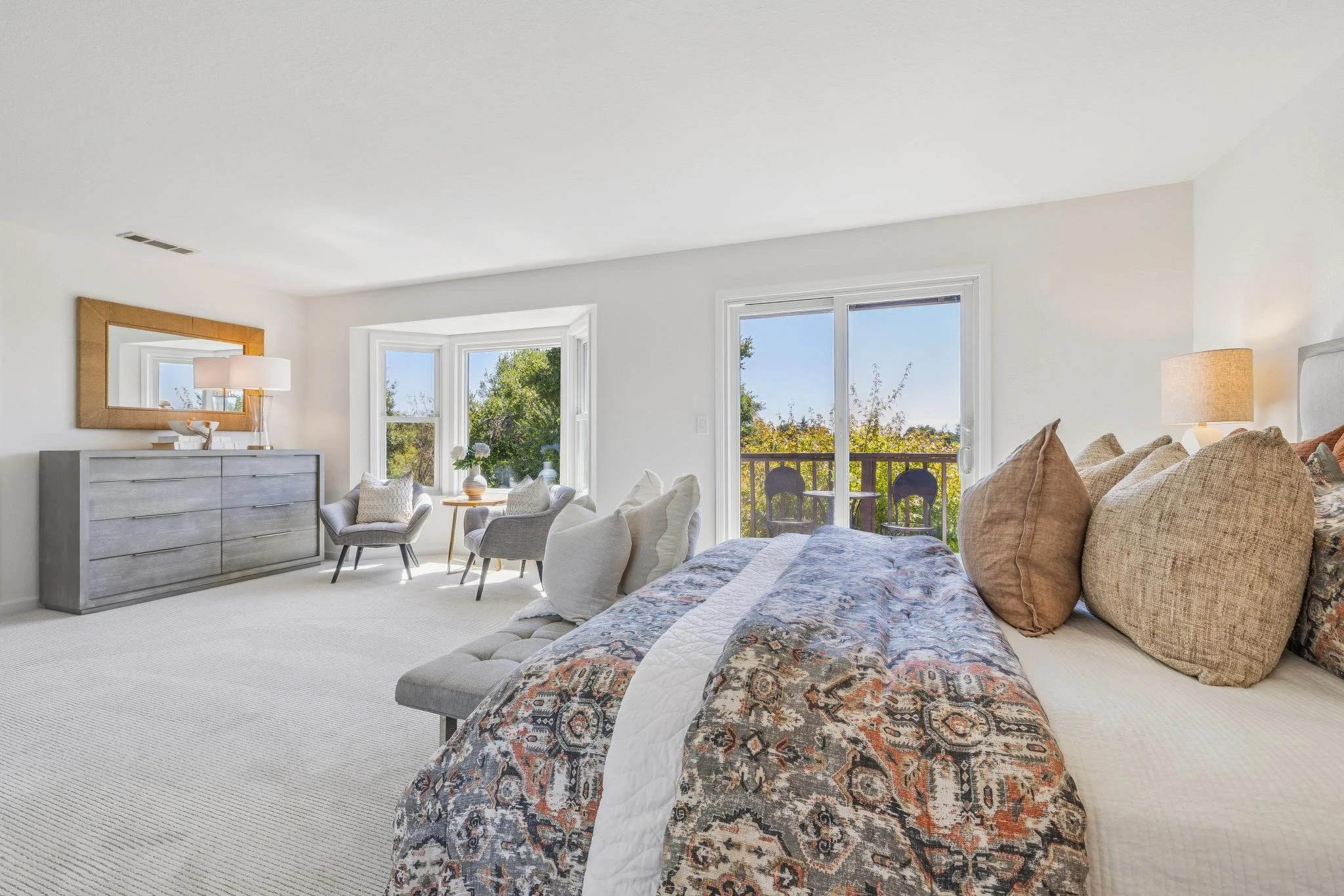 Bright living room with large windows, beige carpet, a gray dresser with a mirror, gray chairs, a floral-patterned bed or sofa with beige pillows, and a sliding glass door leading to a balcony with trees outside.