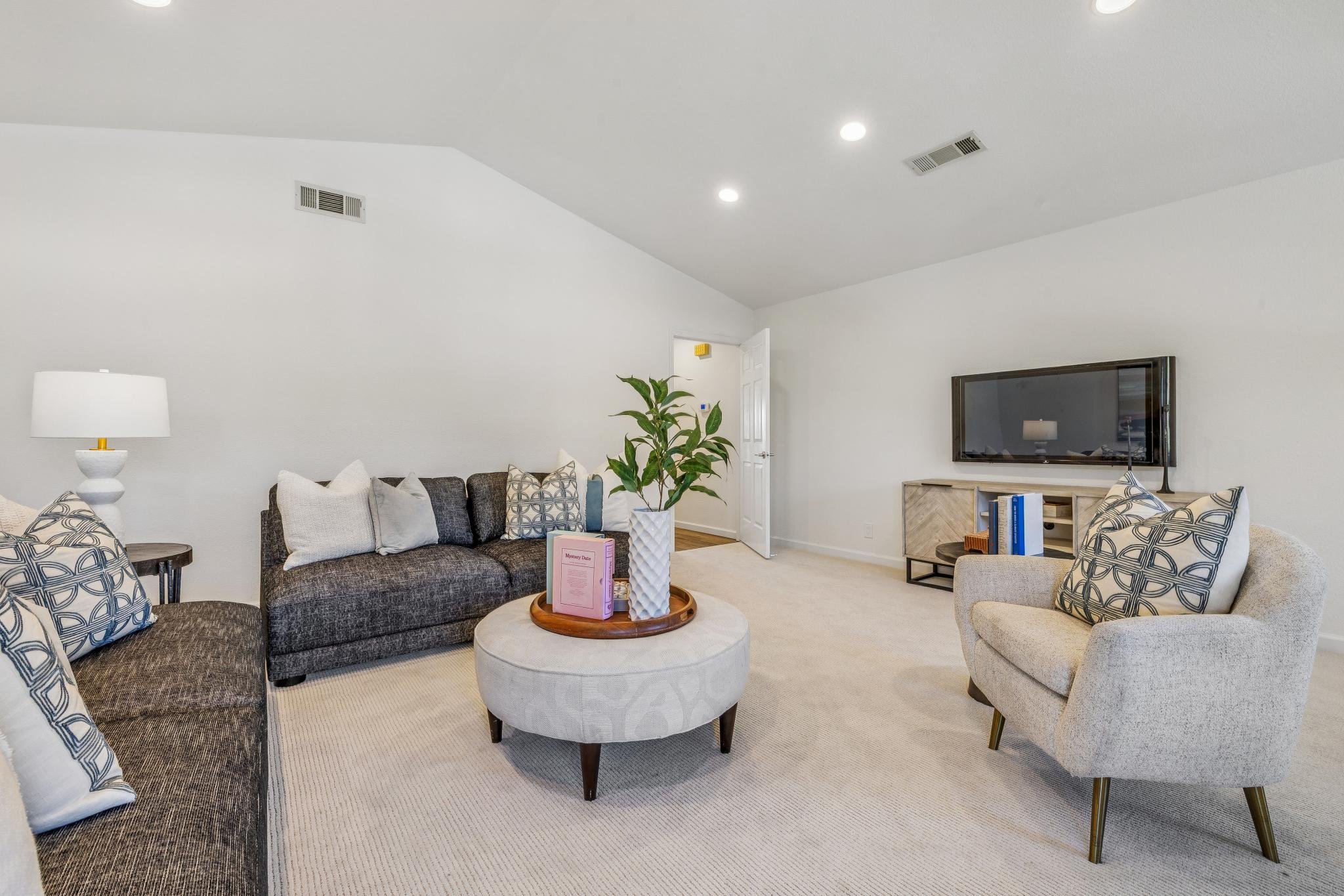Living room with a dark gray sofa, light-colored armchair, patterned pillows, white side table with a lamp, coffee table with a plant and books, wall-mounted TV, and a door in the background.