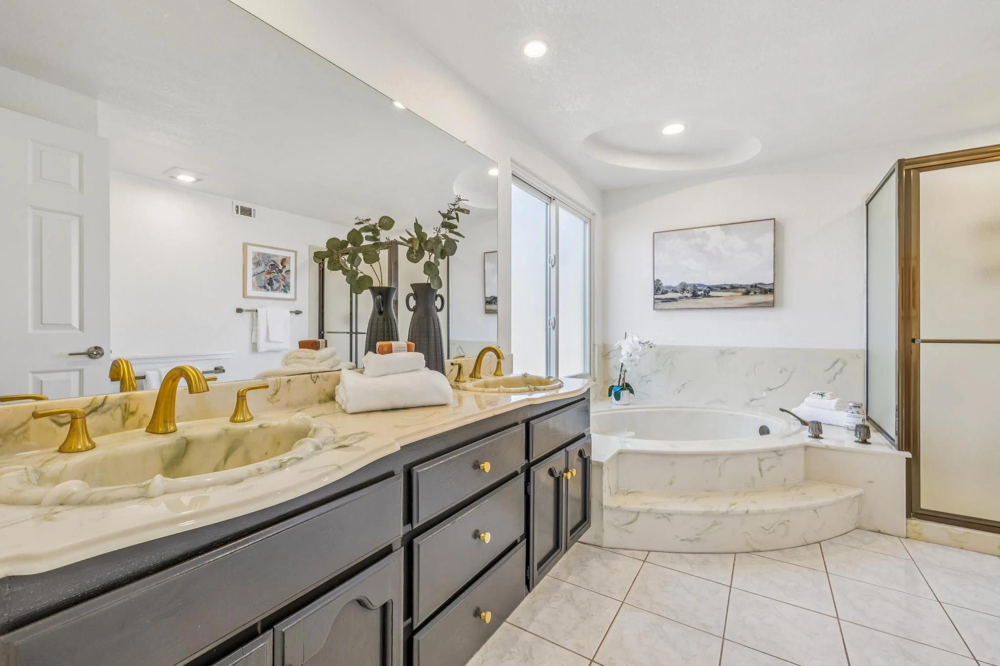 A luxurious bathroom with a dark wood vanity with gold hardware, cream marble countertops, two sinks with gold fixtures, a large mirror above, a bathtub with a marble surround, framed artwork, and decorative vases with greenery.