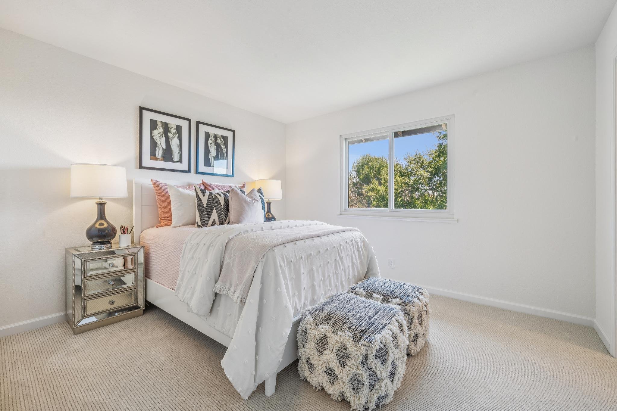 Bright bedroom with white walls and beige carpet. Bed with white bedding and decorative pillows. Mirrored bedside table with a lamp and accessories. Two framed artworks above the bed. Large window showing green trees outside.