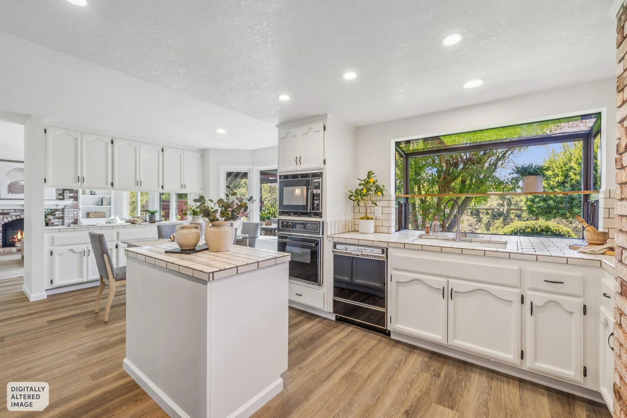 Bright kitchen with white cabinets, a large window with a view of trees, a kitchen island with ceramic tile top, and a stone accent wall.