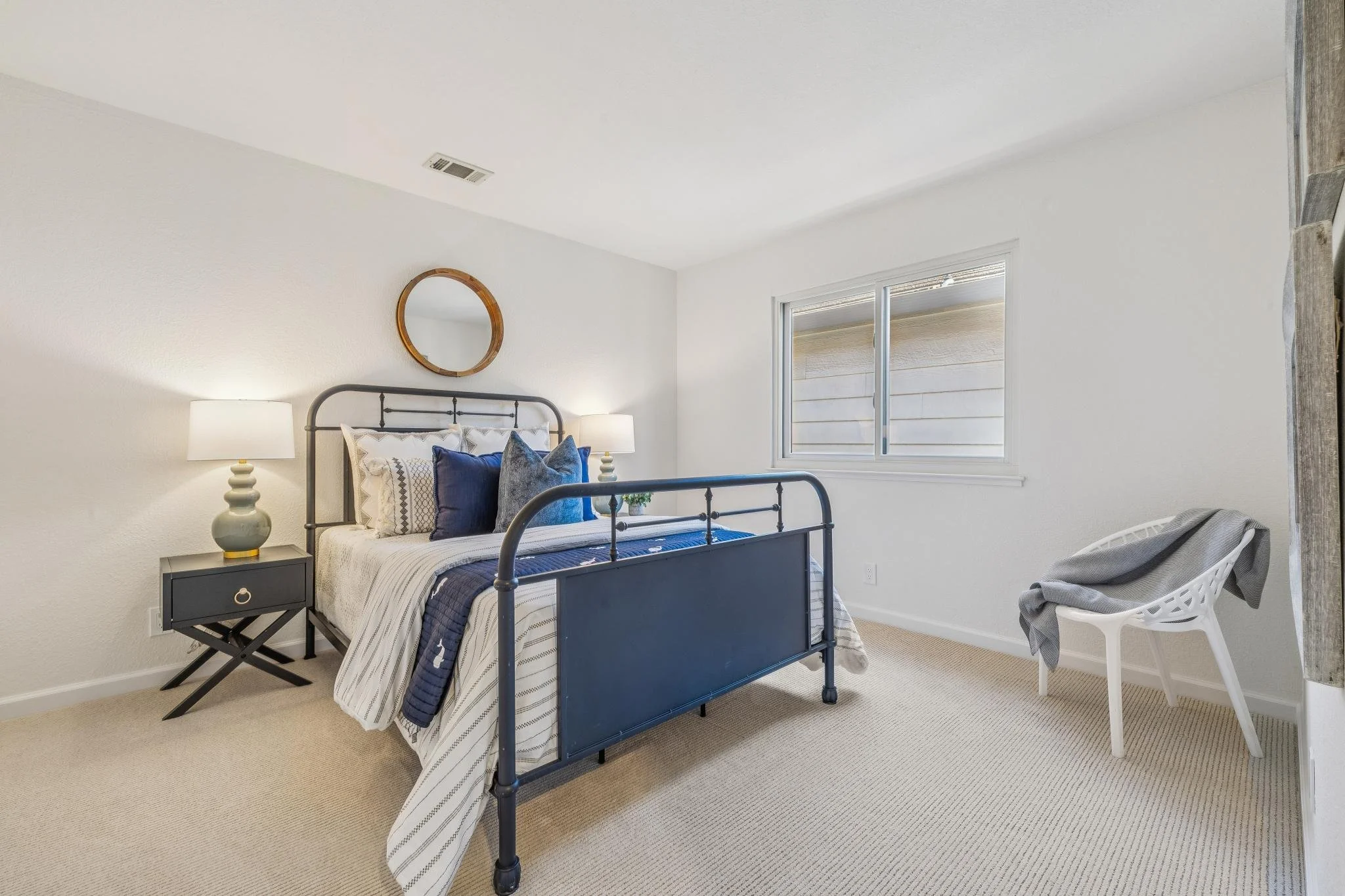 A bedroom with a metal bed frame, white and blue bedding, a nightstand with a lamp, and a chair with a grey blanket.
