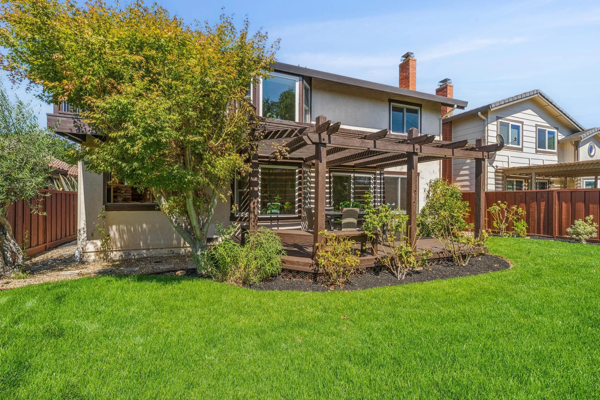 Backyard with green lawn, trees, a wooden deck with a pergola, and neighboring houses in the background.