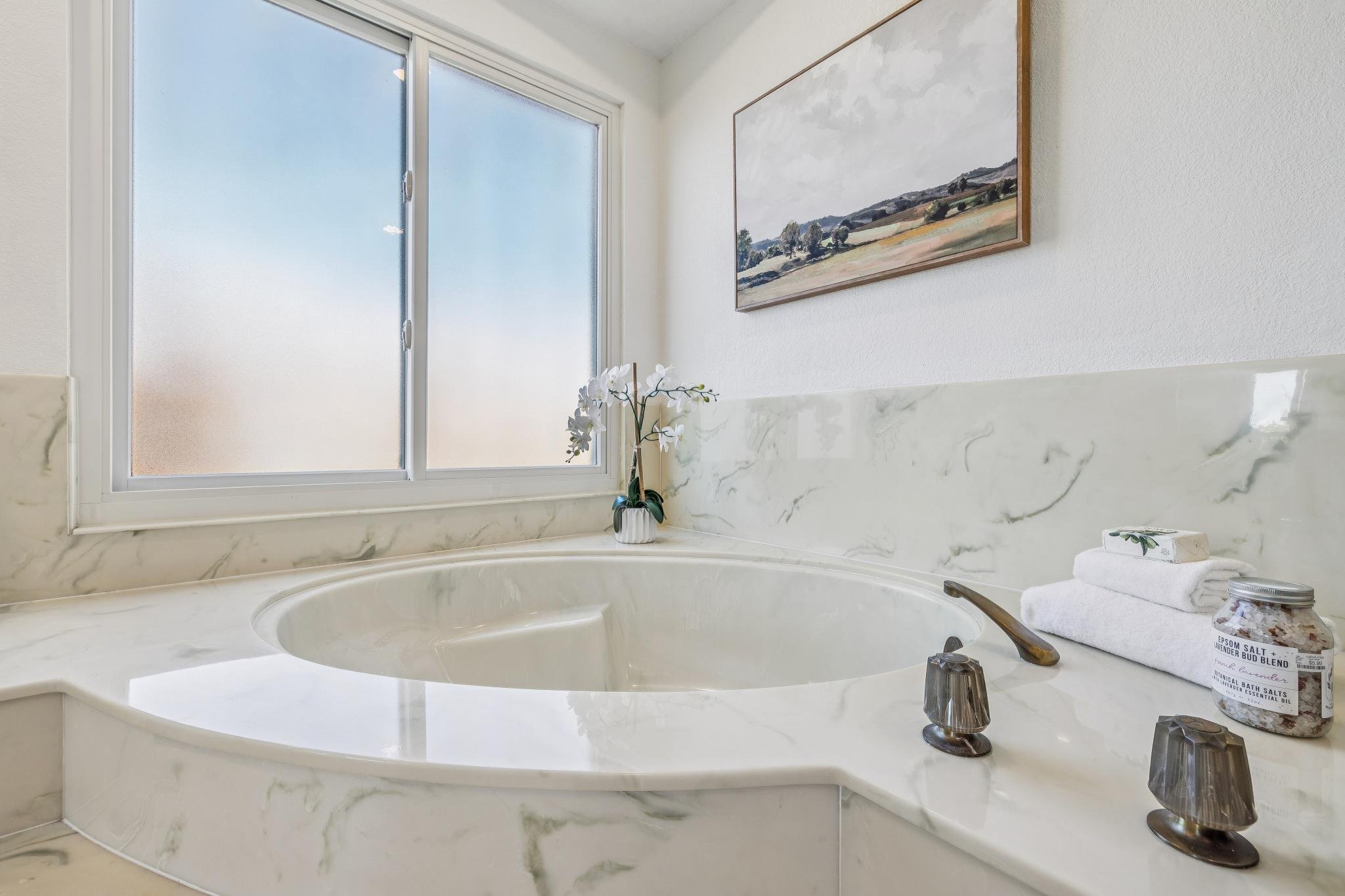 A bathroom with a large frosted window above a marble-topped bathtub. There is a potted orchid, a towel, and bath salts on the edge of the tub, with a painting of a landscape on the wall.