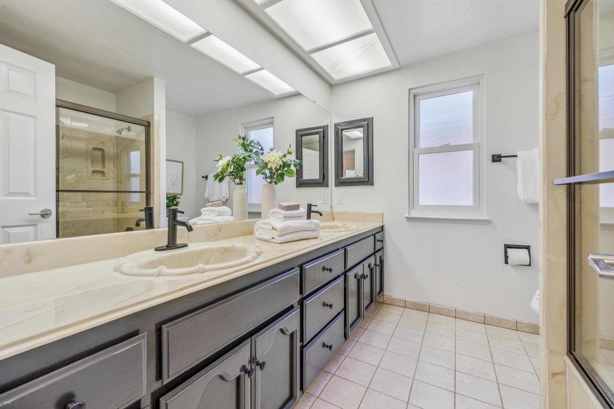 A bathroom with a large marble countertop, dark wood cabinet drawers, a double sink, black faucets, a mirror, and a vase with white flowers. There are windows and a shower with a glass door.