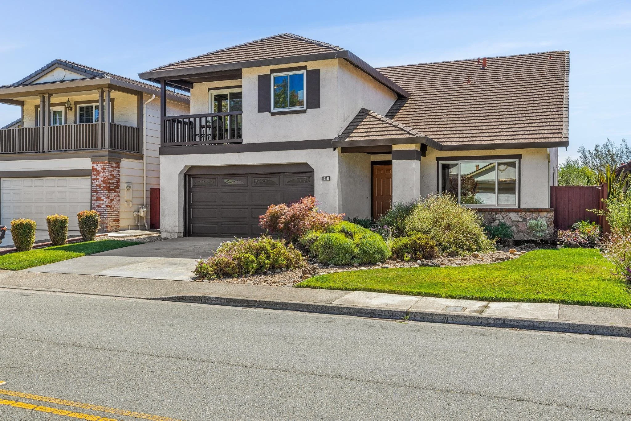 Front view of two neighboring houses with front yards, one with a brown garage door and the other with a balcony, a driveway, and green lawn with bushes and flowers.