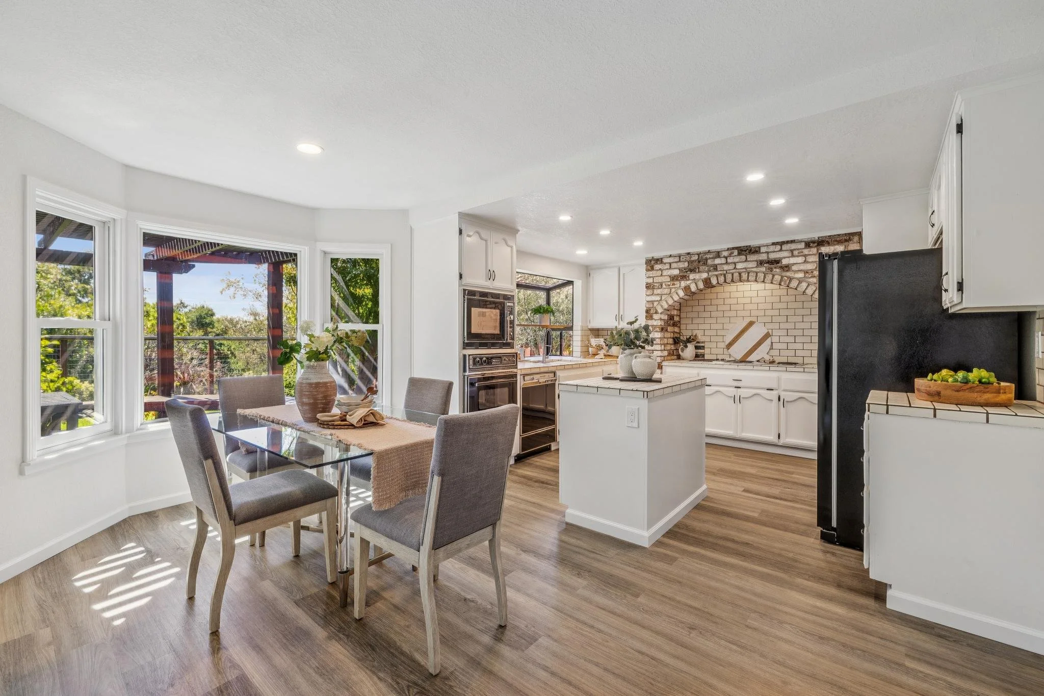 Bright kitchen with white cabinets, brick arch, black refrigerator, dining table with four chairs, large windows, and hardwood flooring.