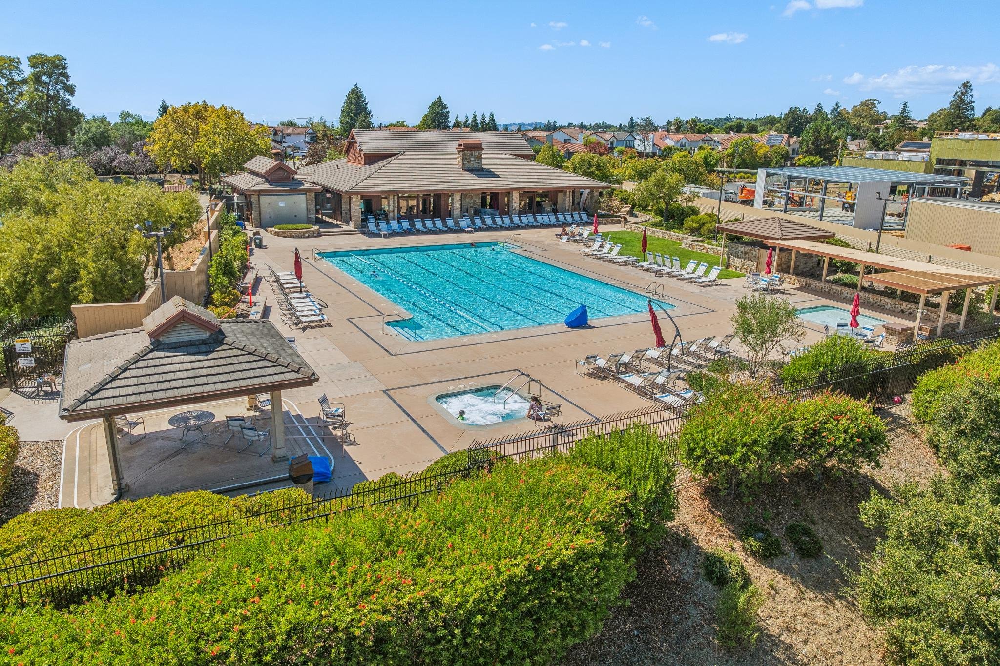 Aerial view of a community swimming pool area with lounge chairs, umbrellas, and a hot tub, surrounded by trees and residential houses.