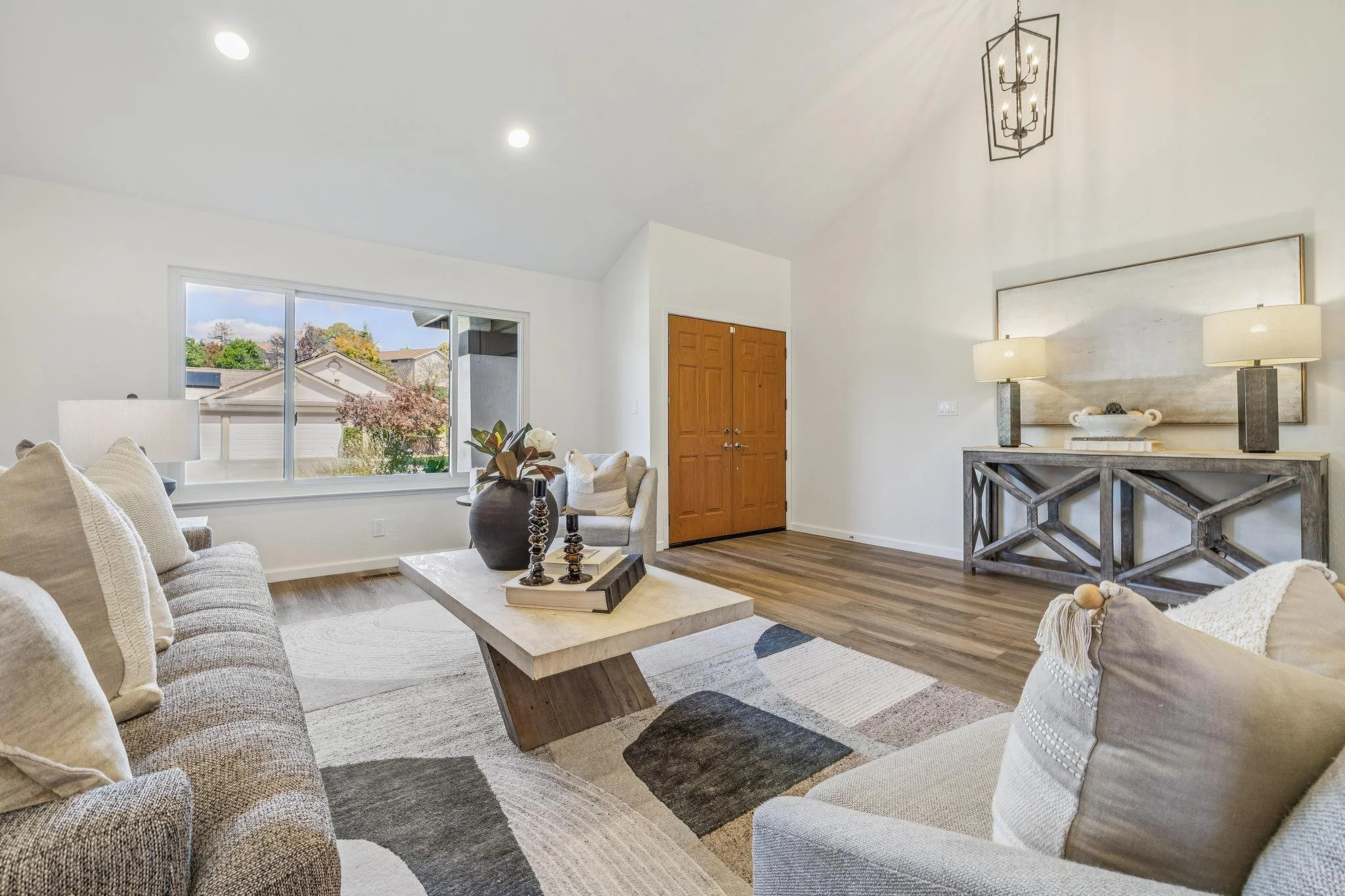 Living room with beige sofas, a white coffee table with decorative items, a large window with a view of trees and neighboring houses, a wooden front door, and a console table with lamps and artwork.