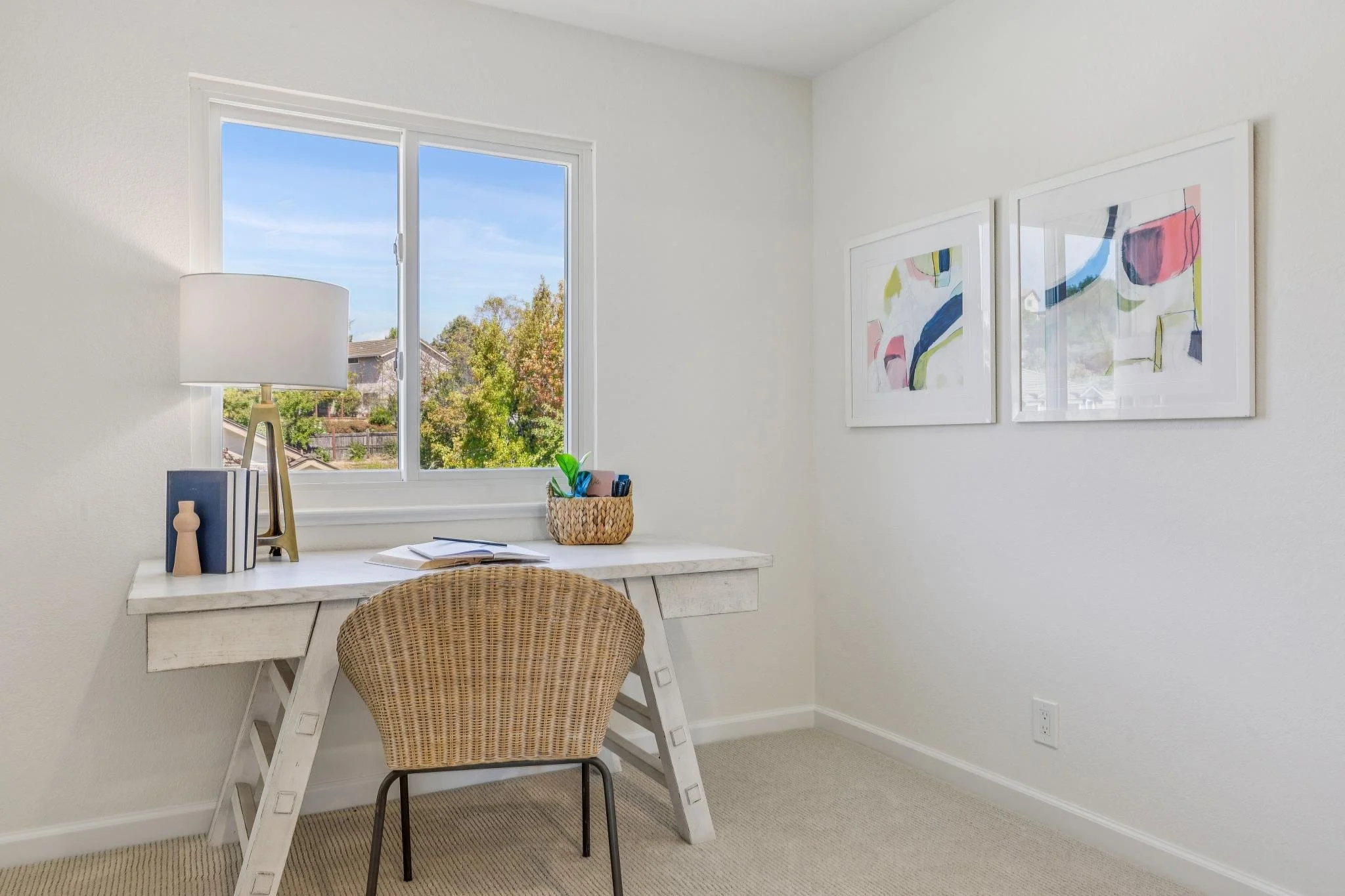 A small home office with a white desk, a wicker chair, and wall art. There is a window showing trees outside, and a table lamp and some books and decorative items on the desk.