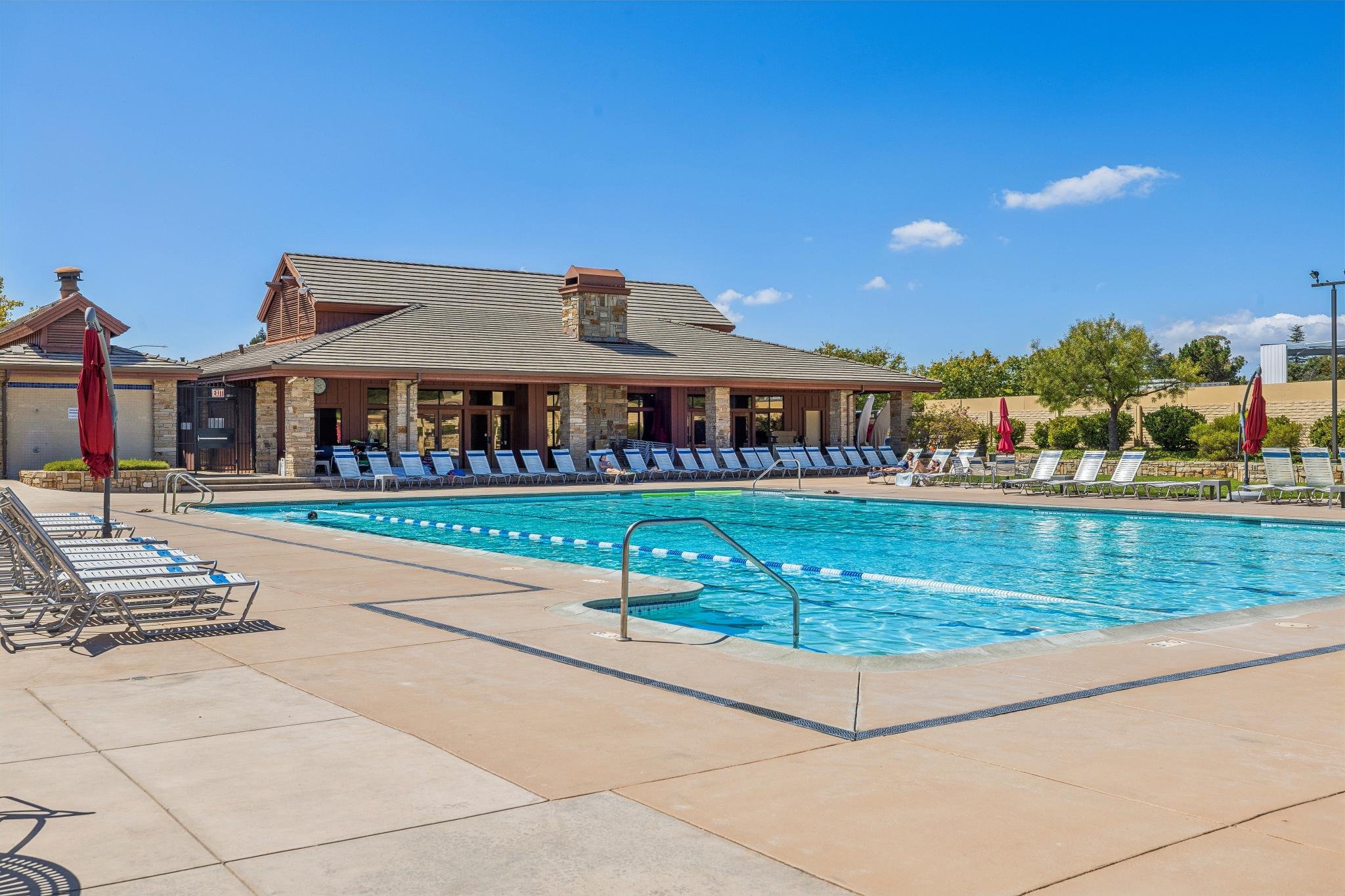 Empty swimming pool with lounge chairs, umbrellas, and a clubhouse building under a blue sky with a few clouds.