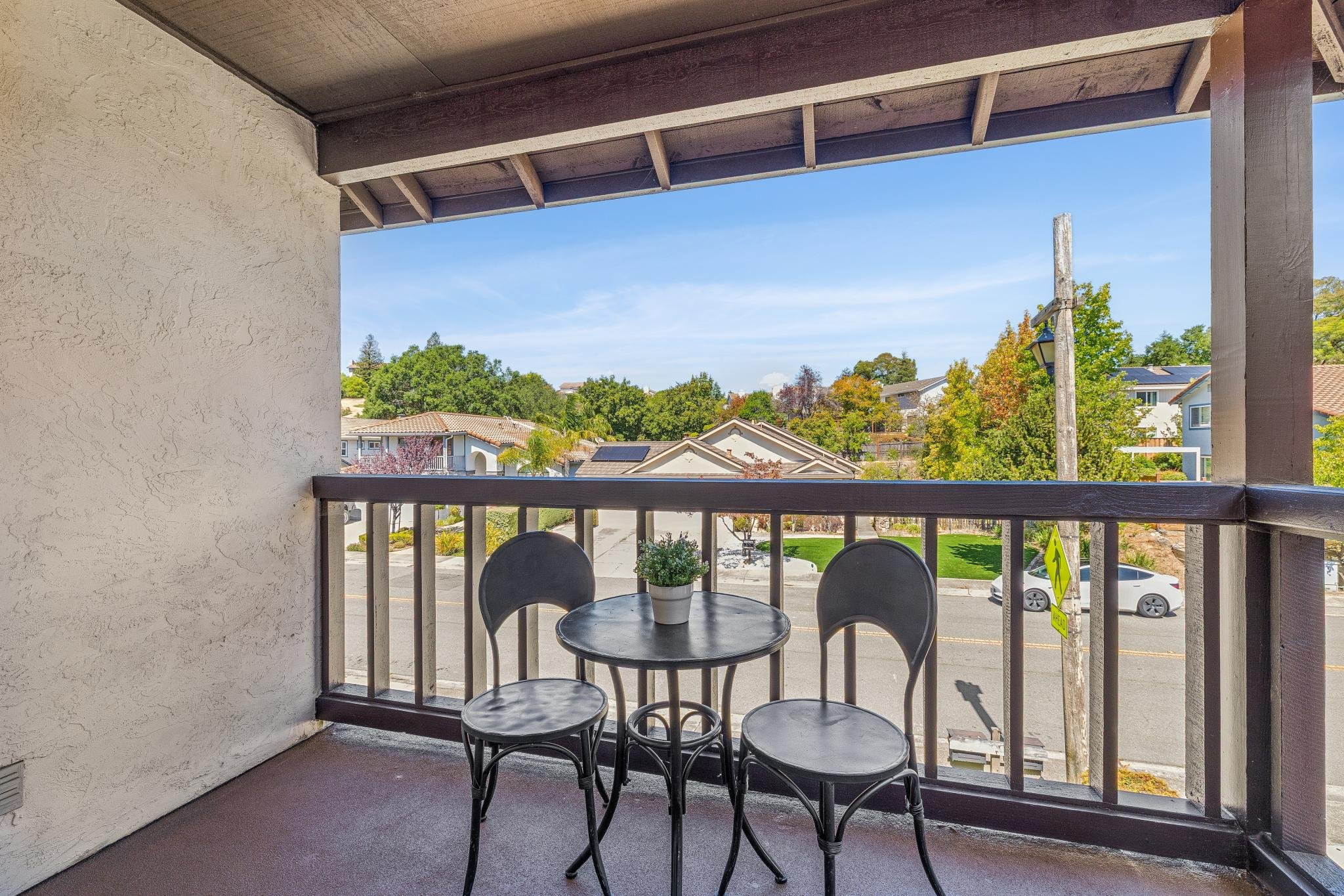 Balcony with three black metal chairs and a round table with a small potted plant, overlooking a residential neighborhood with trees and houses, under a wooden ceiling.