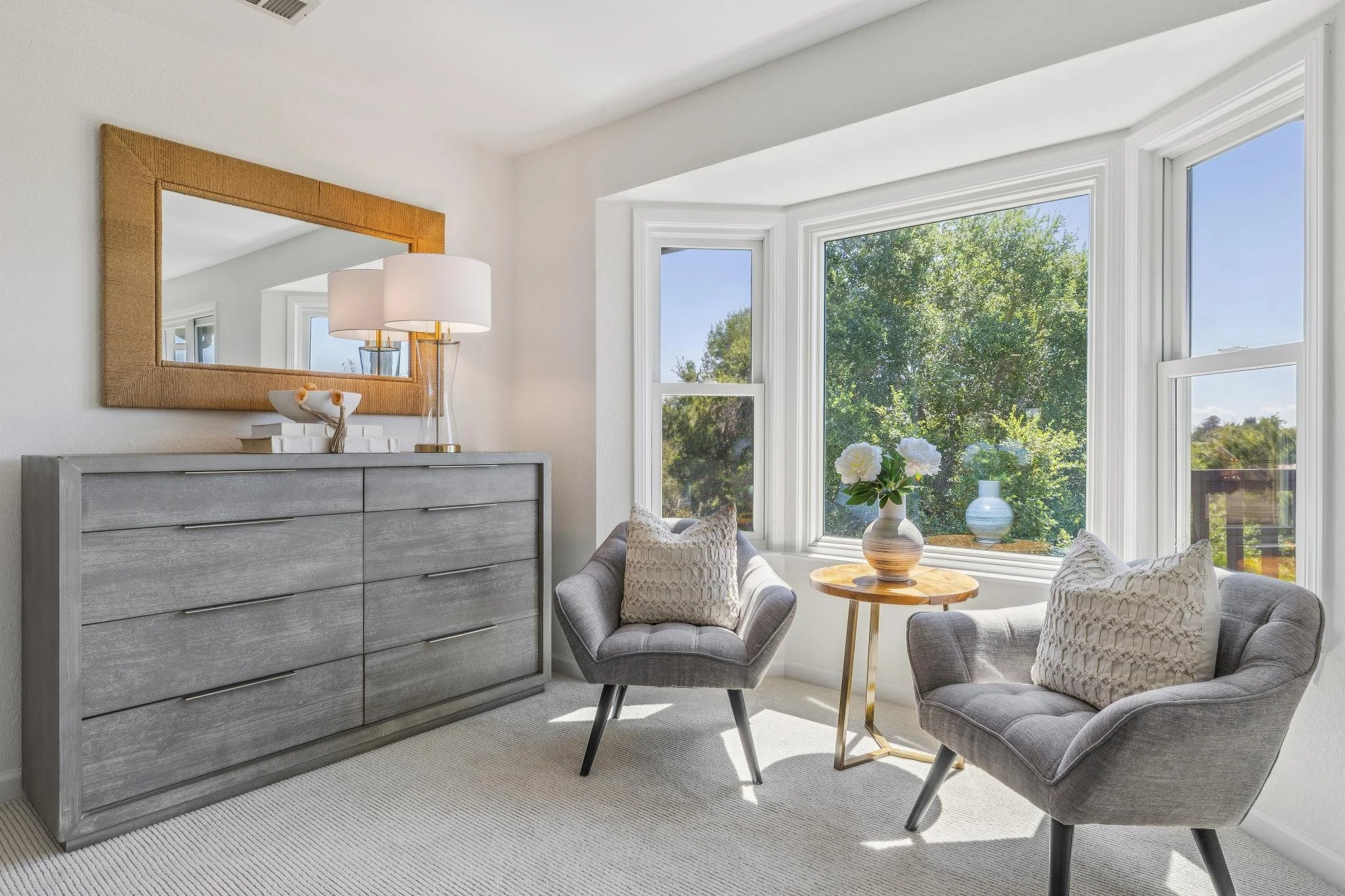 Bright living room with large windows, gray dresser, table lamp, two gray armchairs with decorative pillows, and vases with flowers on a side table.