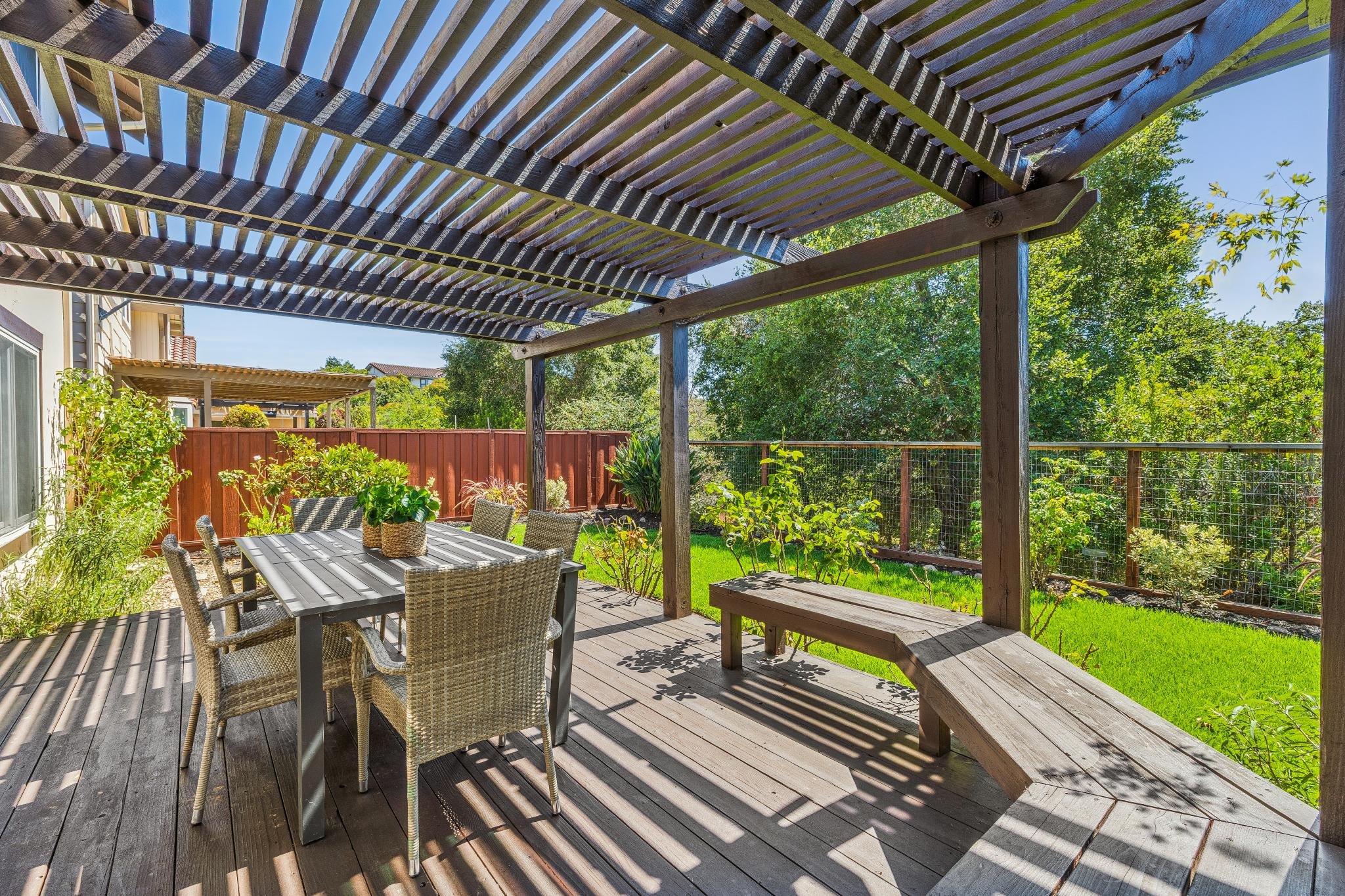 A backyard patio with a wooden pergola, an outdoor dining table with chairs, and lush green trees and grass in the background.