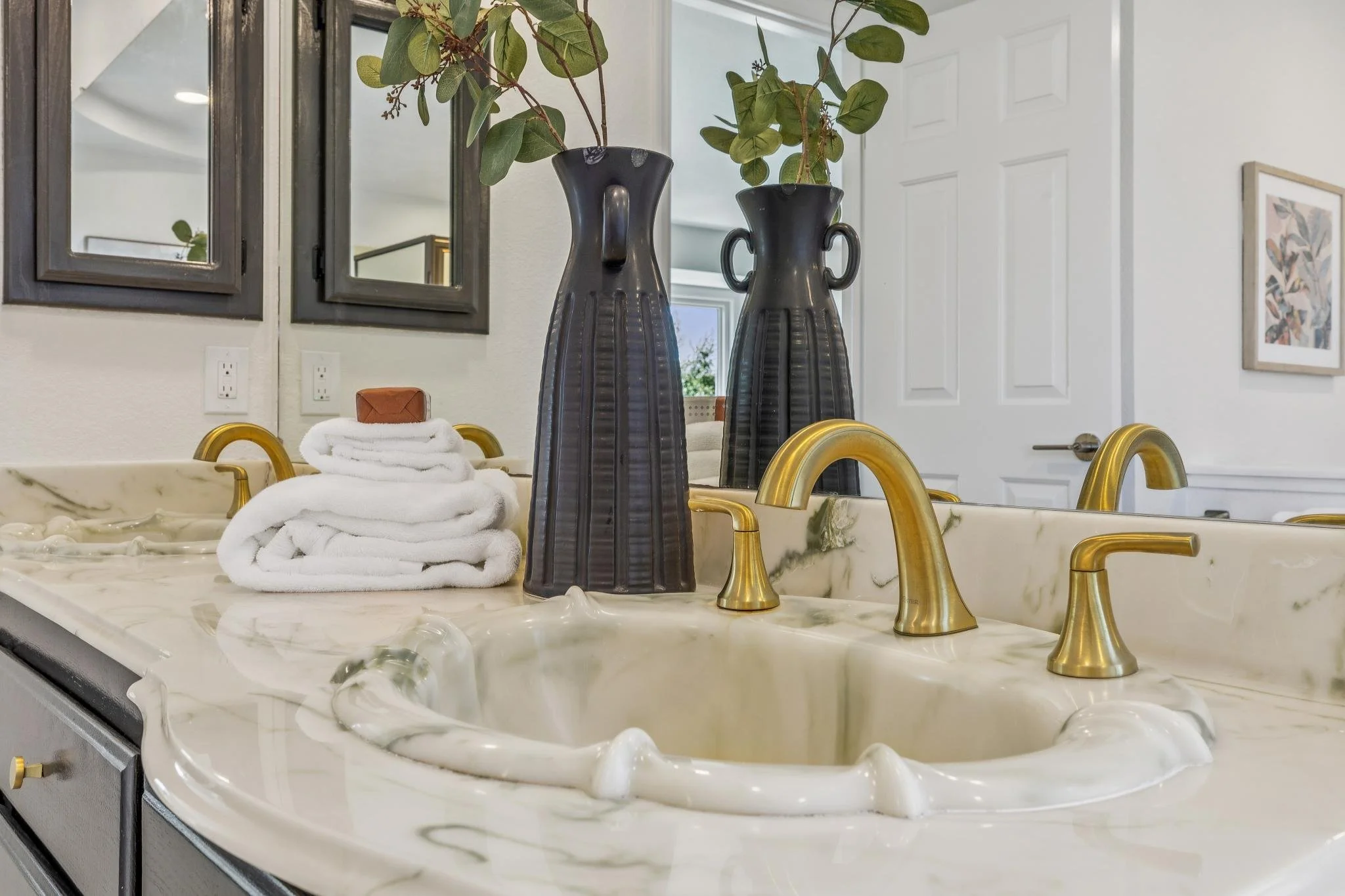 Decorative bathroom countertop with two black vases with green leaves, folded white towels, a bar of soap, and gold-colored faucets, with framed artwork and mirrors in the background.