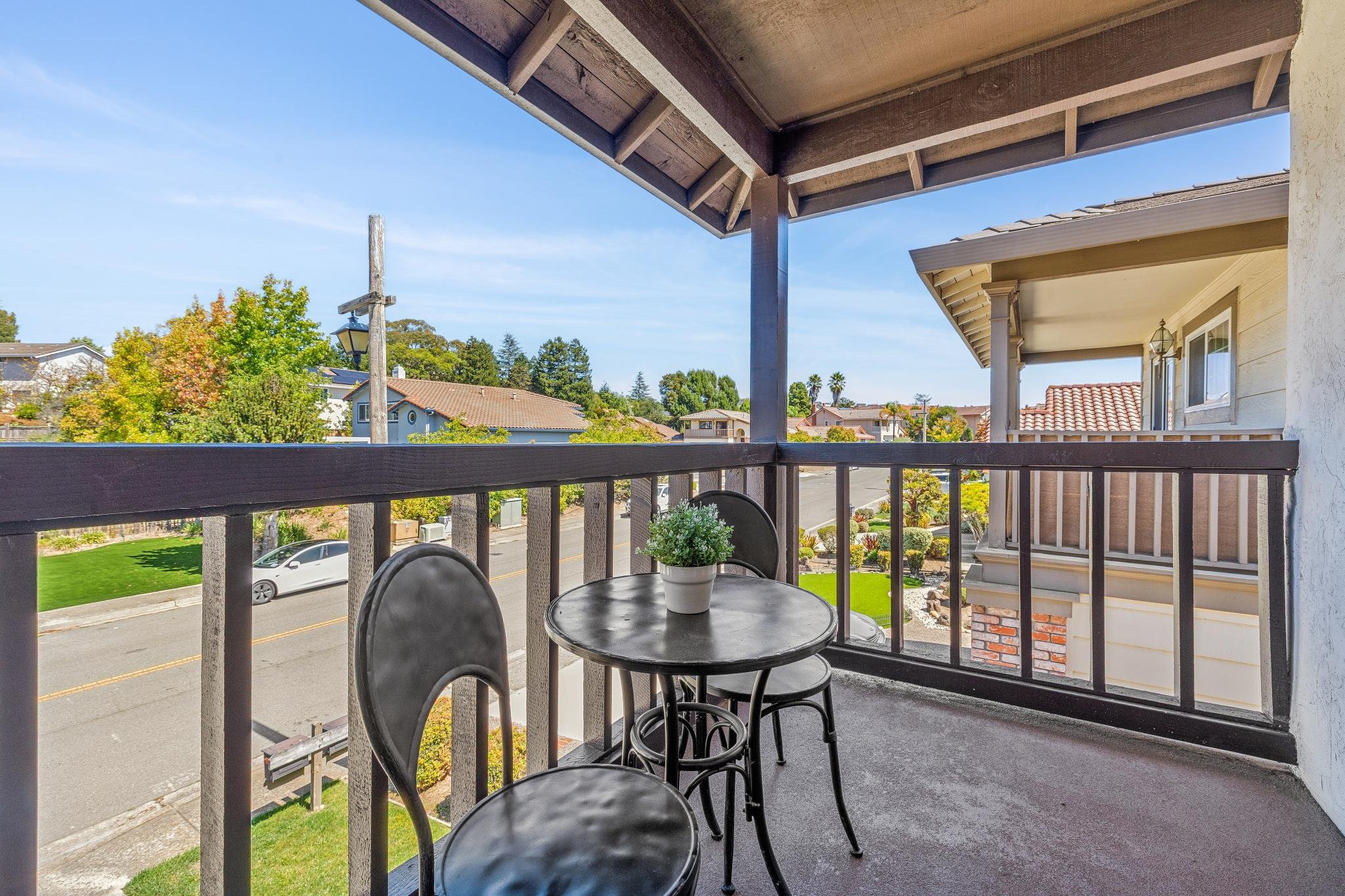 Balcony with a small round table with a potted plant and three chairs, overlooking a suburban neighborhood with trees and houses, under a partly cloudy sky.