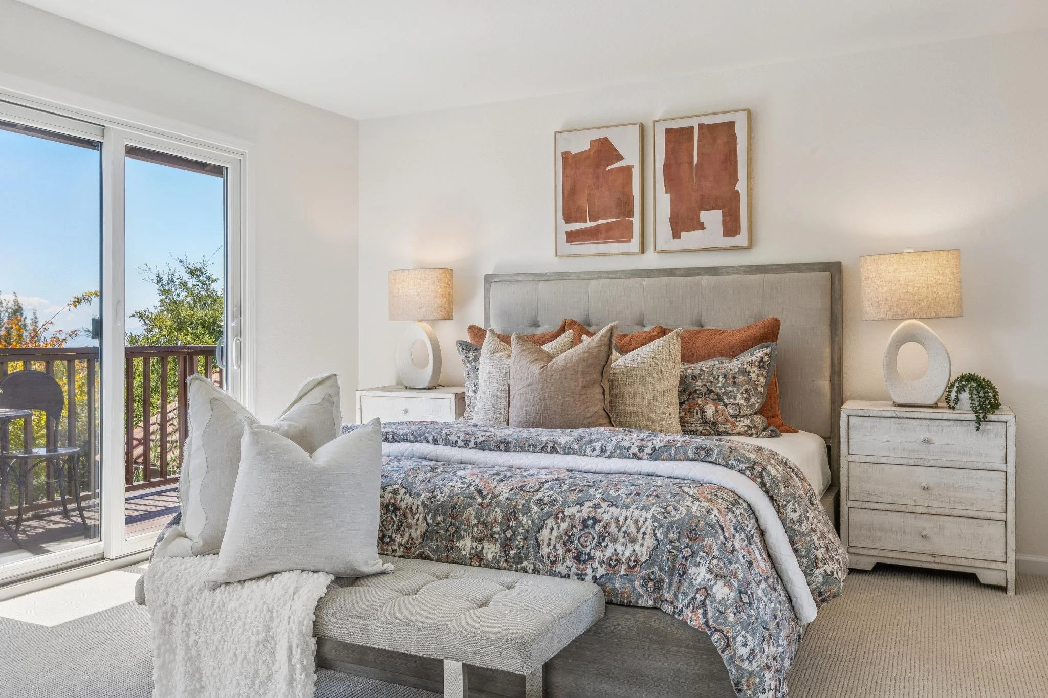 Bright bedroom with a large bed, multiple pillows, a patterned duvet, white furniture, two table lamps, wall art, and a balcony with outdoor furniture and trees visible outside.