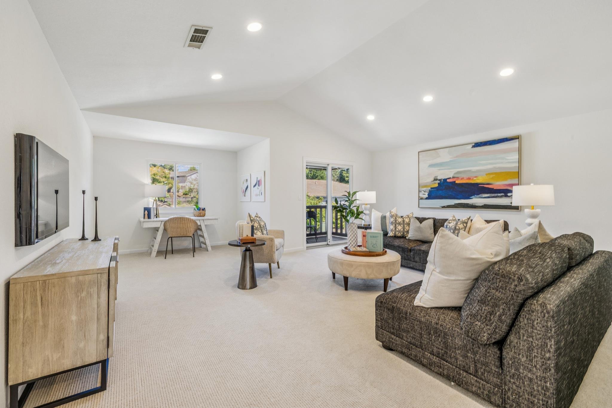 Living room with a beige carpet, dark gray couches, white walls, large colorful abstract painting, white potted plants, white side tables with lamps, sliding glass door leading to a balcony, casual decor, bright and airy atmosphere.
