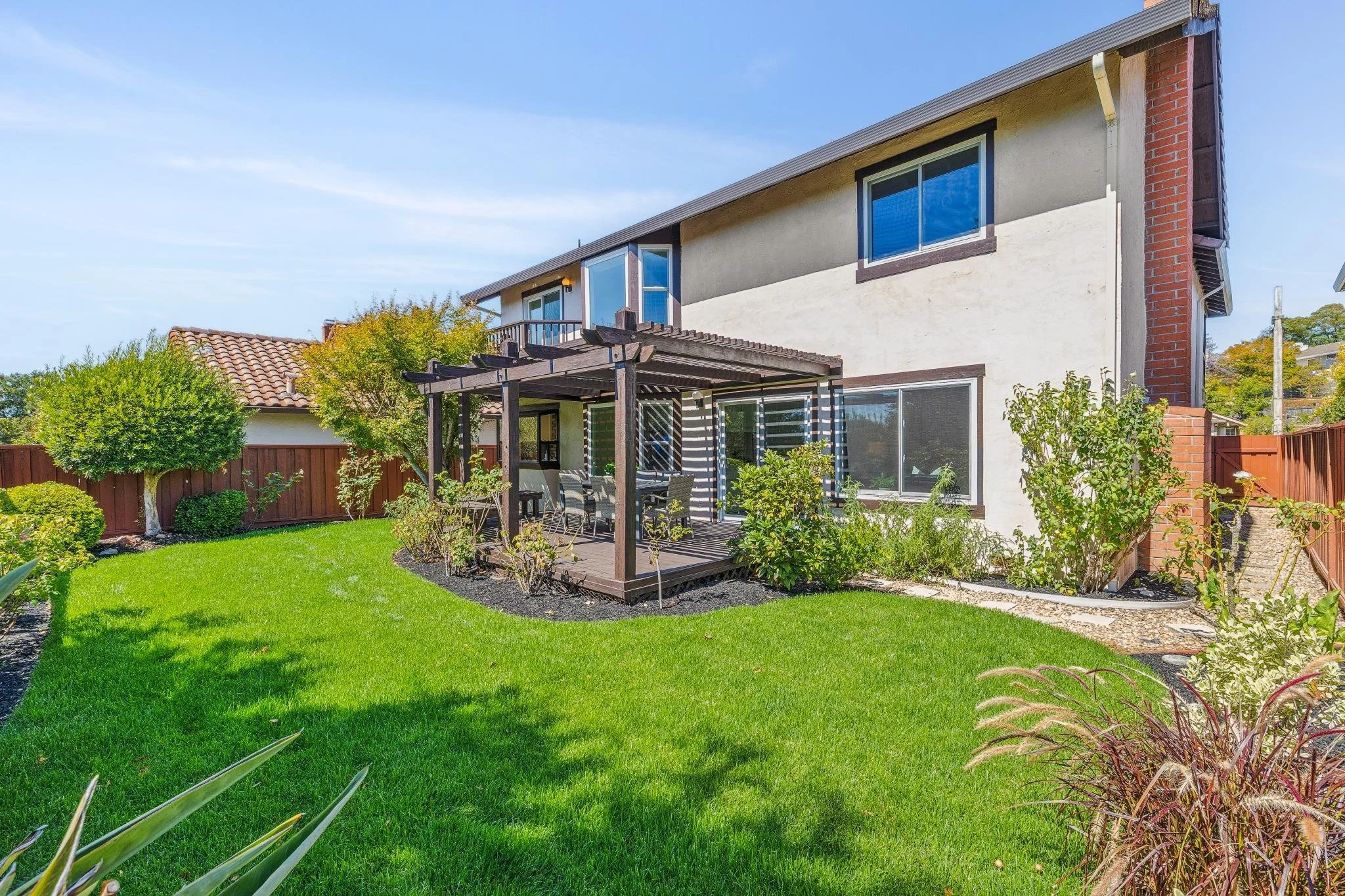 A two-story house with a landscaped backyard featuring a green lawn, a small tree, bushes, and a wooden pergola with outdoor furniture, under a clear blue sky.