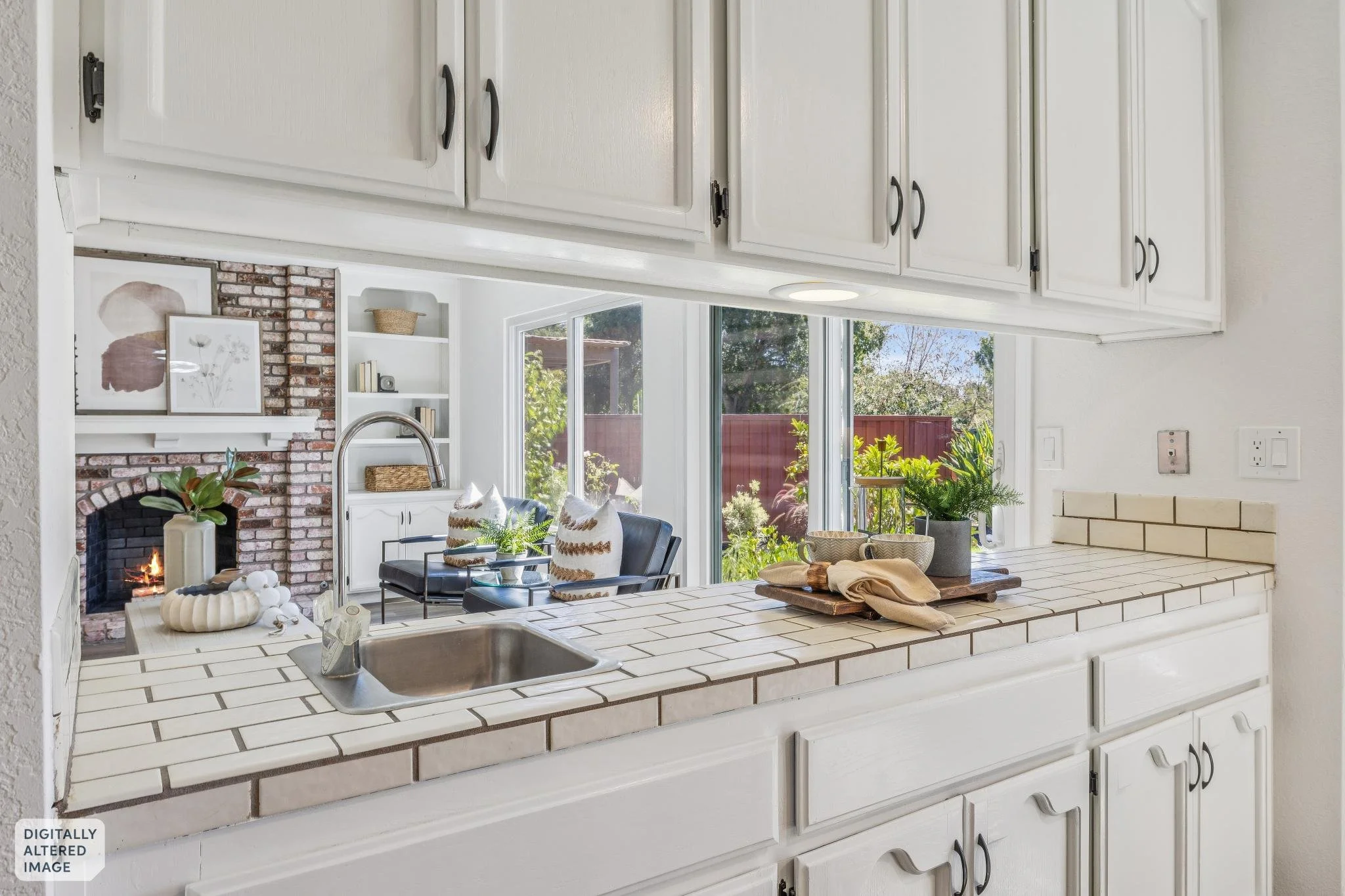 View of a bright, airy living room through a kitchen counter with white tiles. The living room features a brick fireplace with a fire, several framed artworks above it, a white built-in bookshelf, and large windows revealing a garden outside. The roo