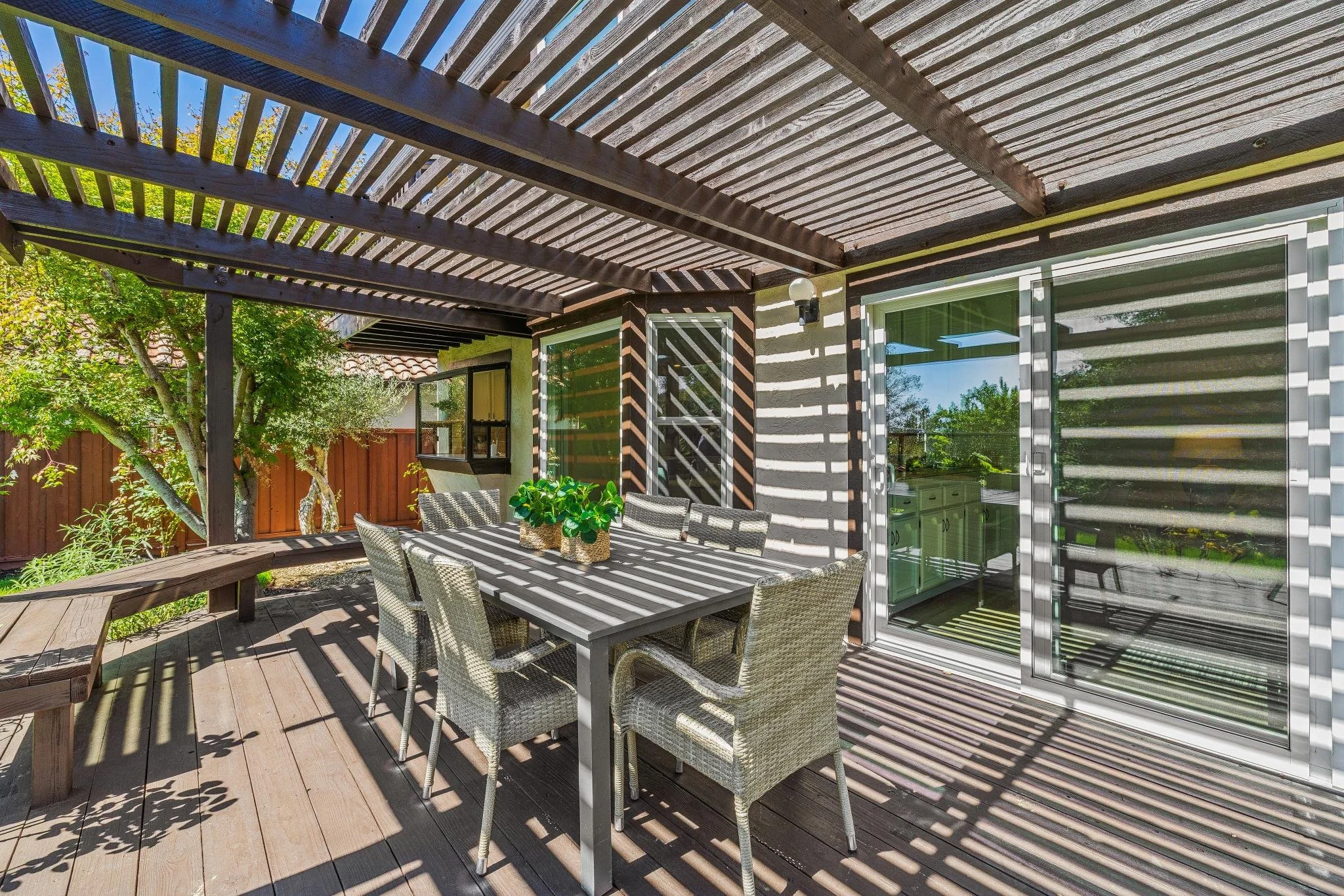 Backyard patio with a wooden table and six chairs under a slatted pergola, with a sliding glass door leading into the house and trees in the background.