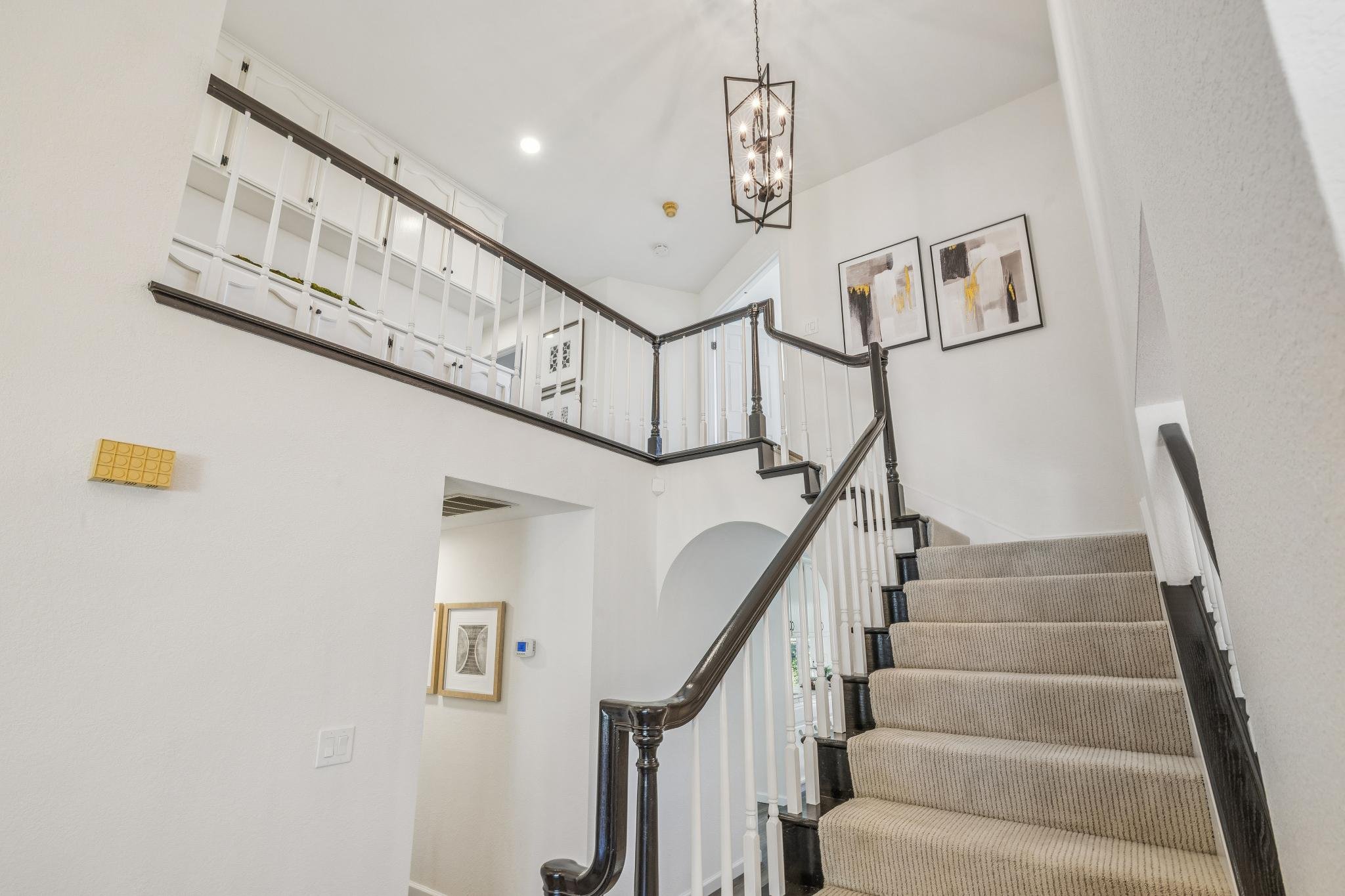Interior view of a staircase with beige carpet, wooden handrail, white balusters, and framed abstract art on the wall. Hanging light fixture with multiple bulbs illuminates the area.