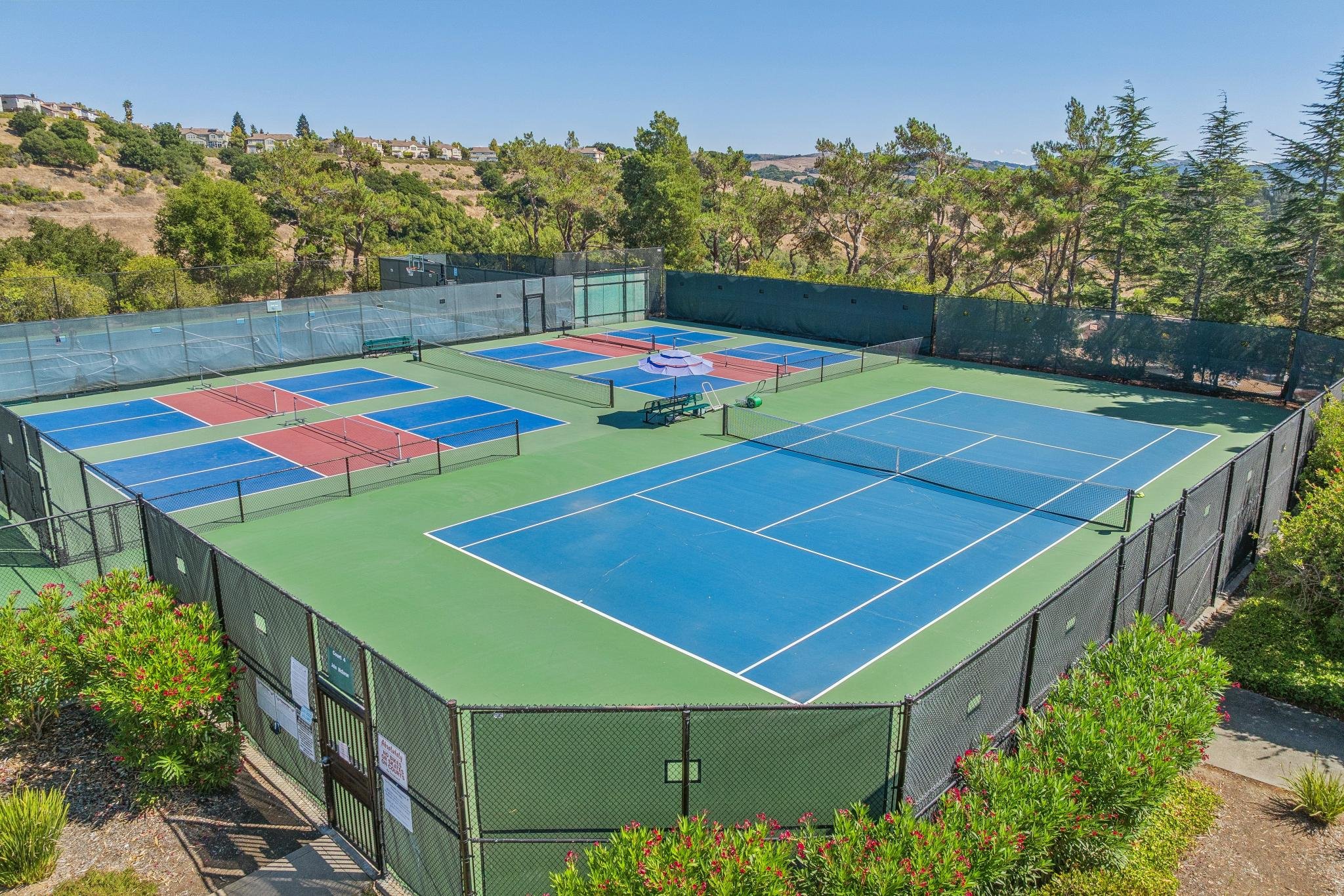 Outdoor tennis courts with colorful blue and red surfaces, surrounded by a black chain-link fence, with trees and hilly residential area in the background under a clear blue sky.