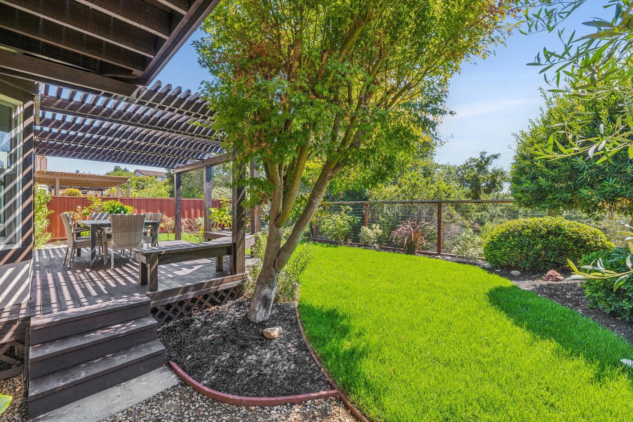 A backyard patio with a wooden deck, outdoor dining table, chairs, and lush green grass with a large tree and plants, under a sunny blue sky.