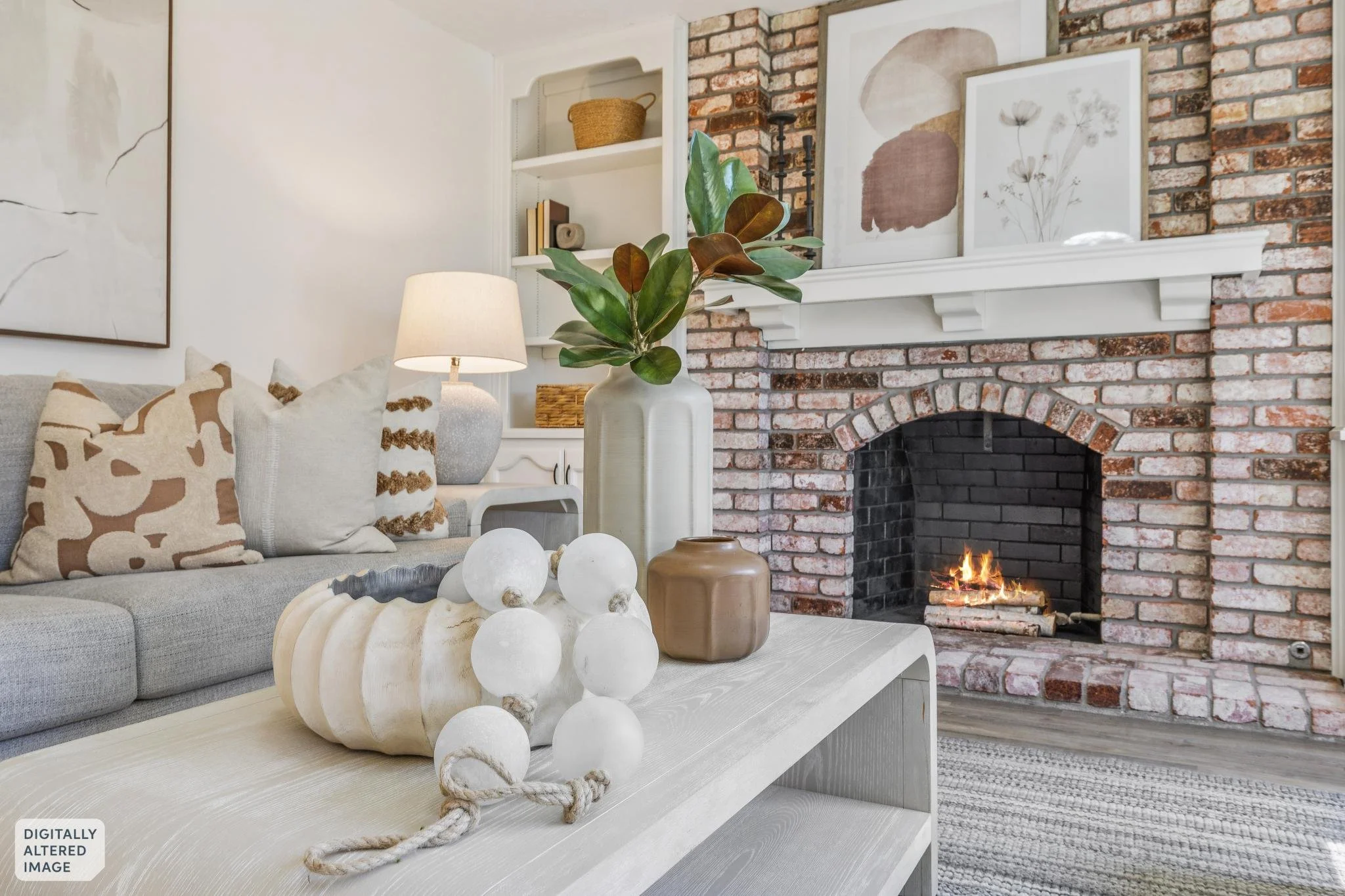 Living room with a sofa decorated with patterned pillows, a table with decorative white orbs, a Murano glass vase with green leaves, a white lamp, and a brick fireplace with artwork above the mantel.