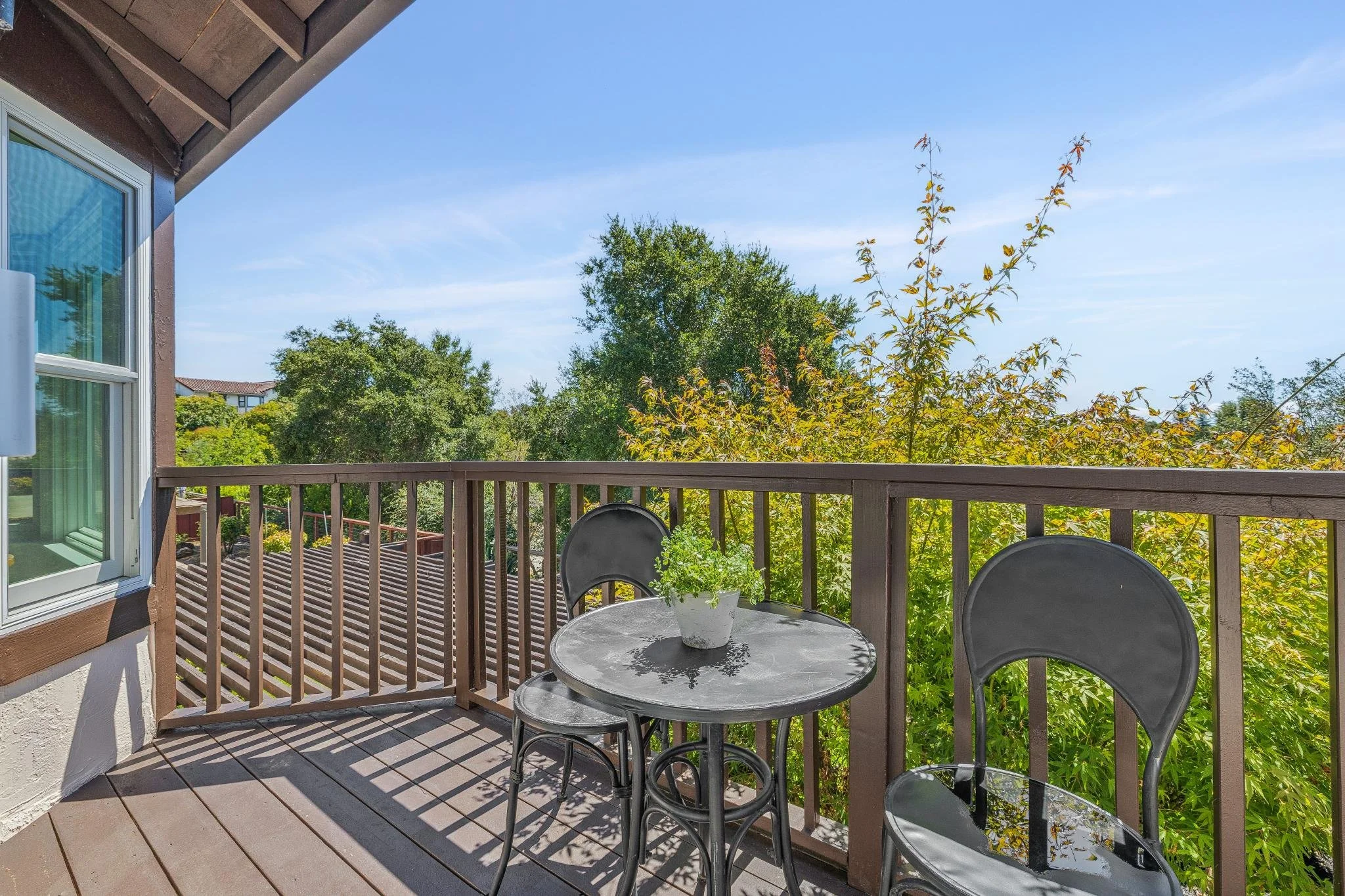 Balcony with a round table and three chairs, potted plant on the table, view of trees and blue sky.