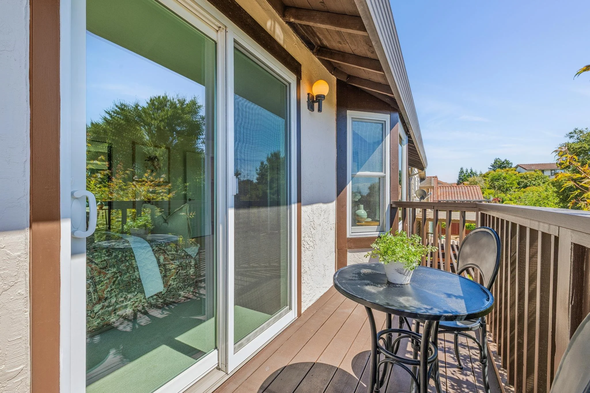 Balcony with black round table, two chairs, potted green plant, sliding glass door, and exterior wall light fixture, with a view of neighboring rooftops and trees under a clear blue sky.