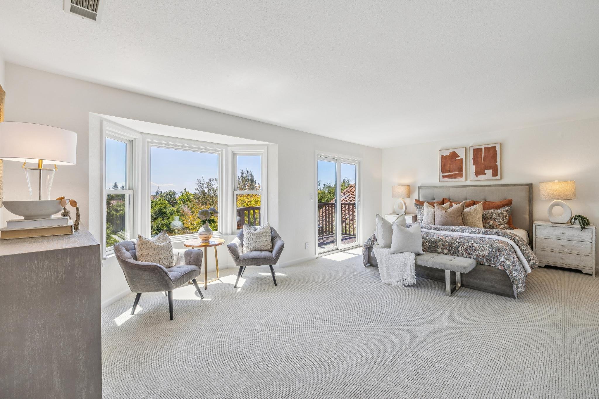 Bright living room with large windows, a bed with pillows, bedside tables, lamps, and framed artwork on the wall.