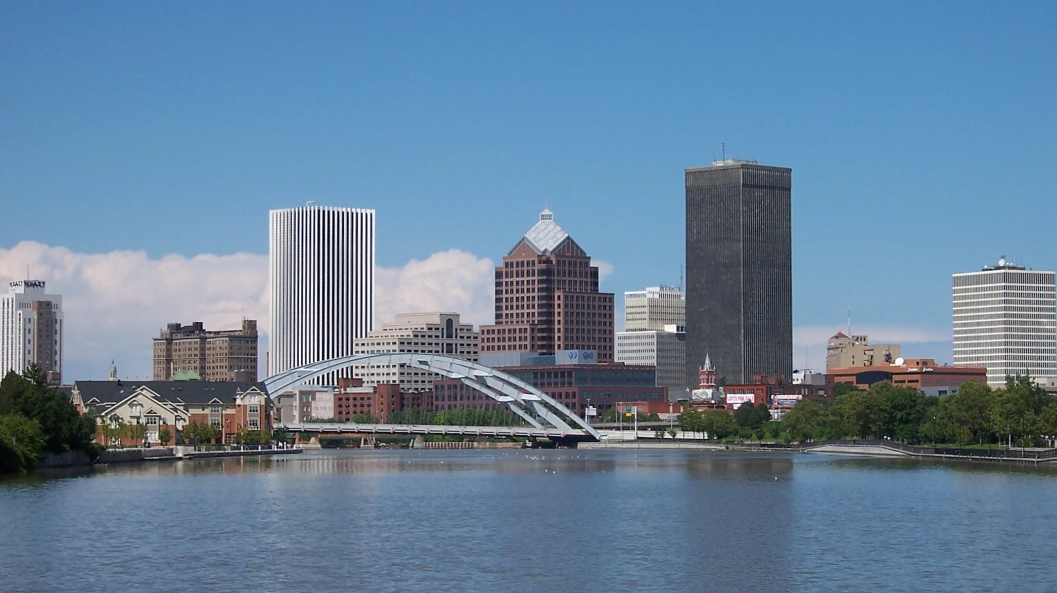 City skyline with tall buildings, a bridge over a river, and a partly cloudy blue sky.