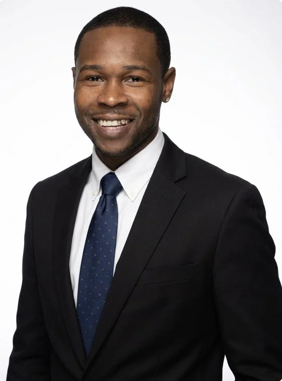 A smiling African American man in a black suit, white shirt, and blue tie, standing against a plain white background.