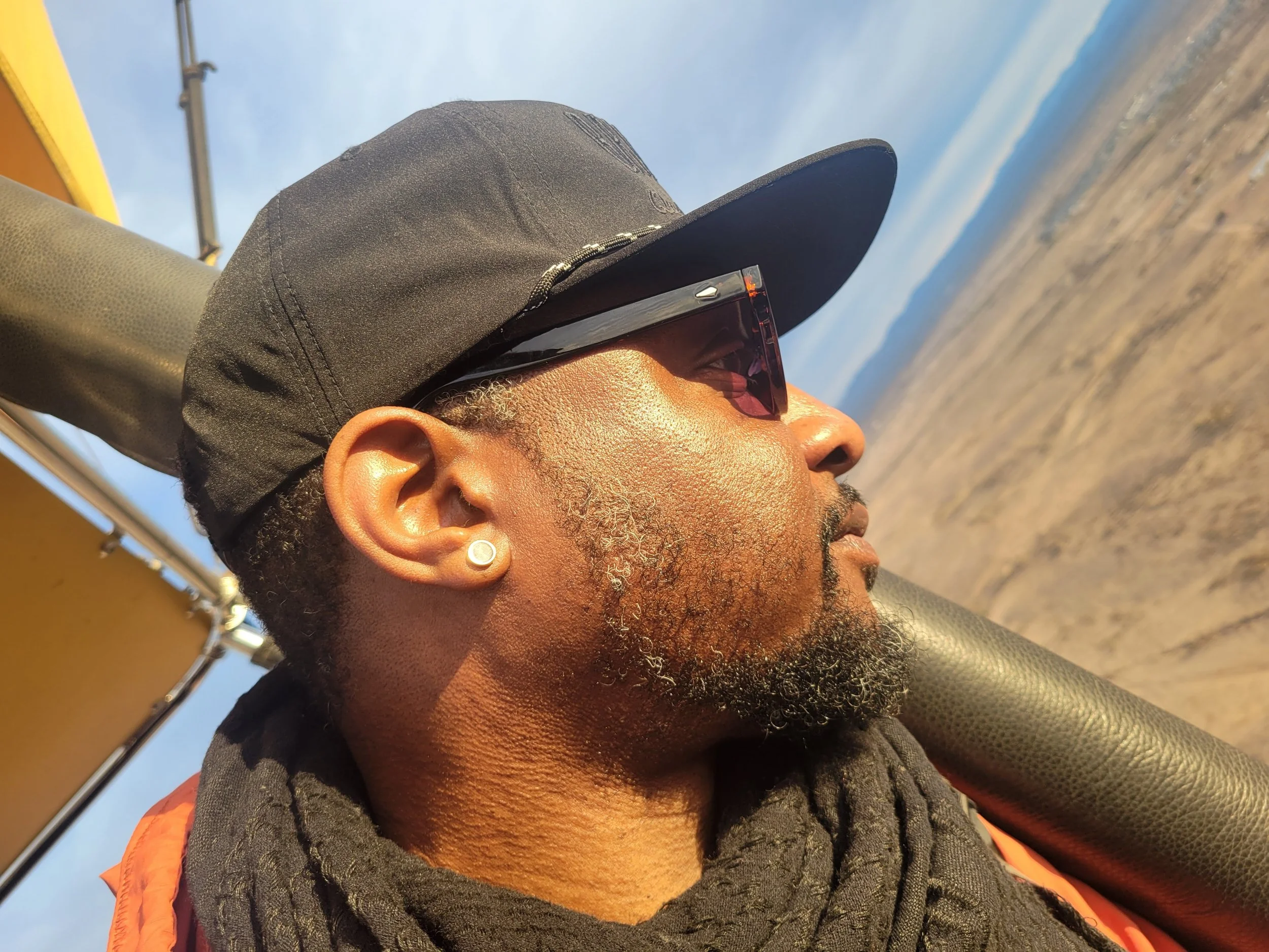 Close-up profile of a man wearing a black cap, sunglasses, and an orange vest, with a landscape of fields and mountains in the background.