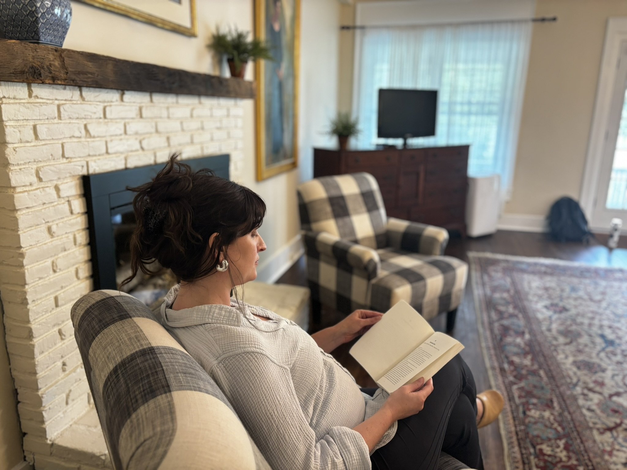 A woman with dark, curly hair reading a book while sitting on a plaid armchair in a cozy living room.