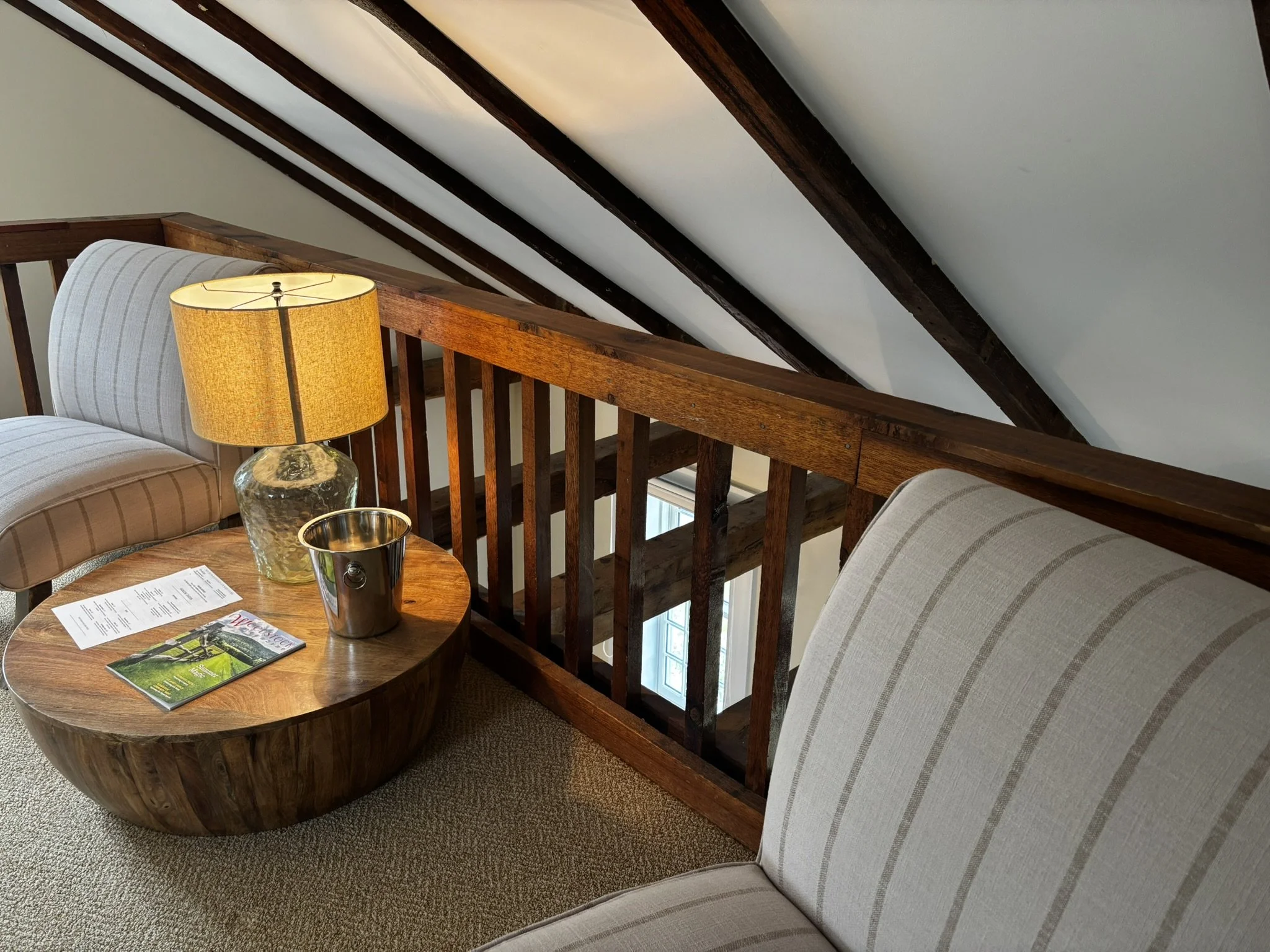 A cozy corner with two beige armchairs with striped patterns, a round wooden coffee table with brochures and a magazine, a glass table lamp with a yellow shade, and wooden ceiling beams.