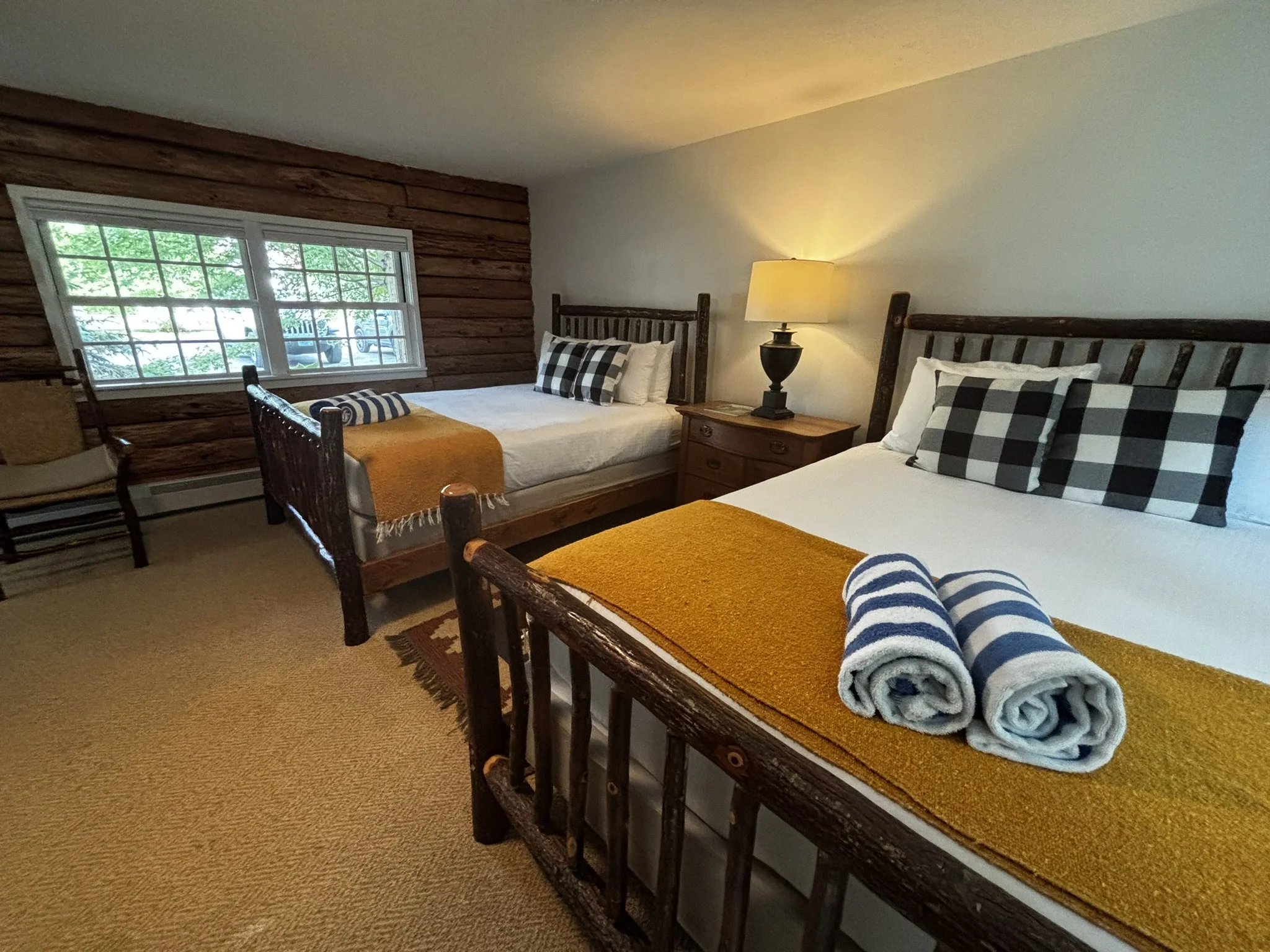 A cozy bedroom with two twin beds made of dark wood. Each bed has a beige blanket, black and white checkered pillows, and rolled towels on top. A bedside table with a lamp is between the beds. A window on the left wall with a view of green trees and a parked car outside.