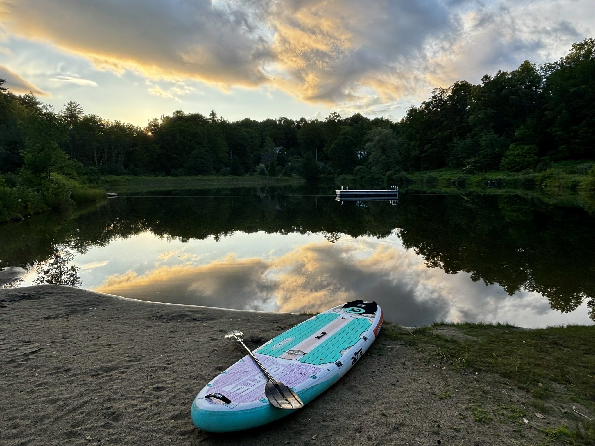 A paddleboard with a paddle resting on a sandy shore near a calm body of water, surrounded by lush green trees, with a dock extending over the water and a partly cloudy sky reflecting on the surface at sunset.