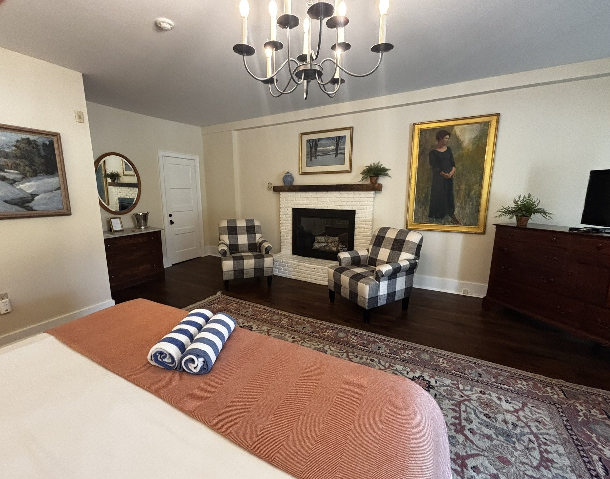 A living room with two plaid armchairs in front of a white fireplace, framed artwork, a mirror, wooden furniture, a chandelier, a potted plant, a TV, and a patterned rug on dark hardwood floors. Towels are on an orange mat.