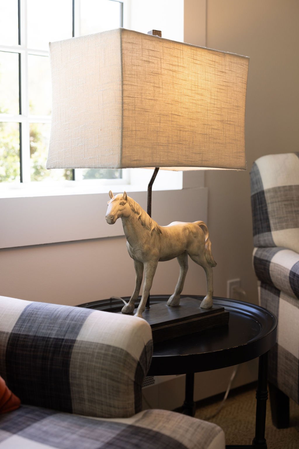 A table lamp with a horse-shaped base and a rectangular fabric lampshade, placed on a black round table between two plaid-patterned chairs near a window.