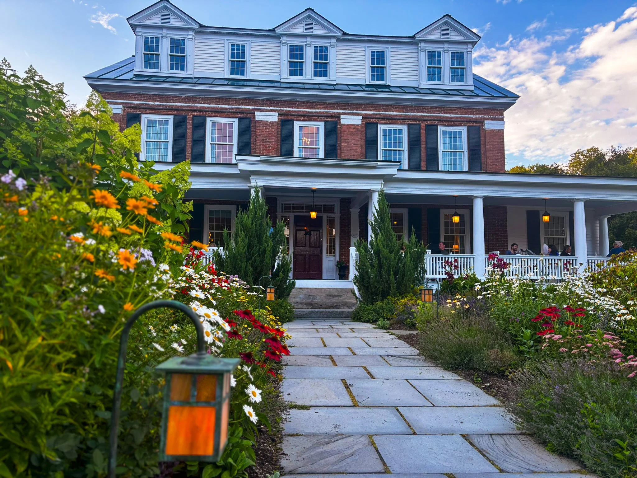 A large brick house with a porch, white columns, and multiple windows, some with black shutters, surrounded by a colorful flower garden and a stone pathway leading to the entrance.