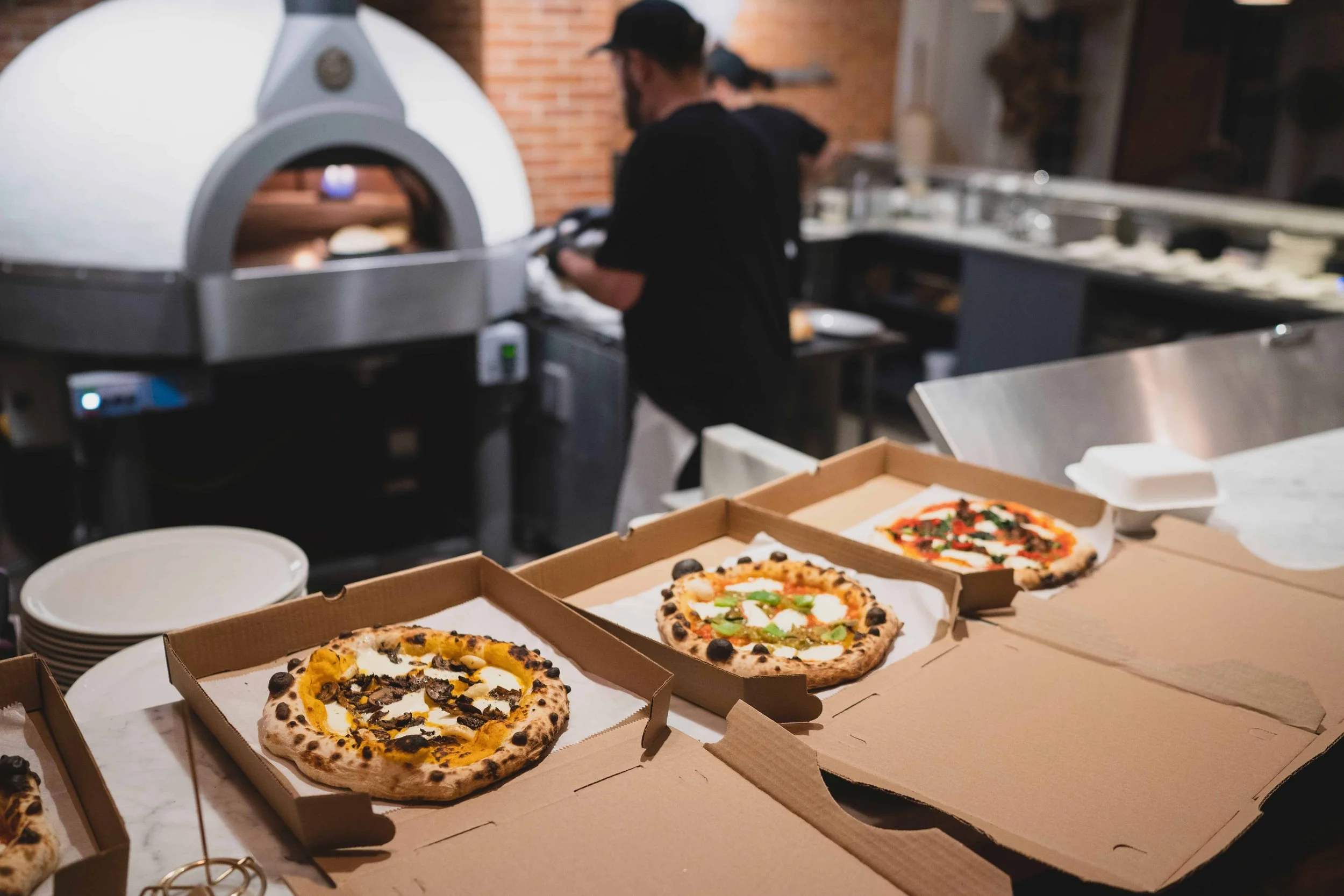 Several pizza slices in open boxes on a table, with a pizza oven and person in background preparing more pizzas in a pizza restaurant.
