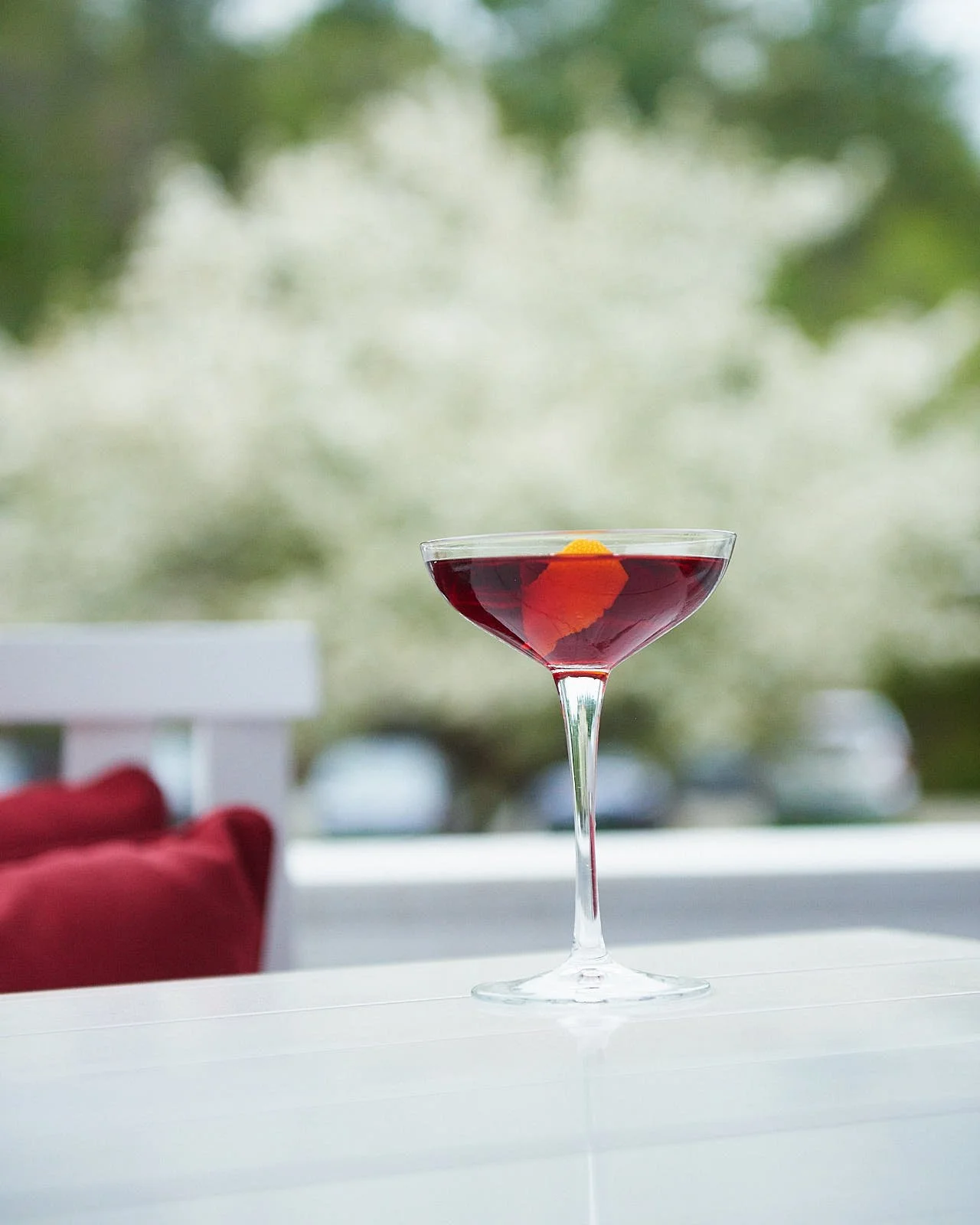 A martini glass filled with red cocktail, garnished with an orange peel, on a white surface with a blurry outdoor background.