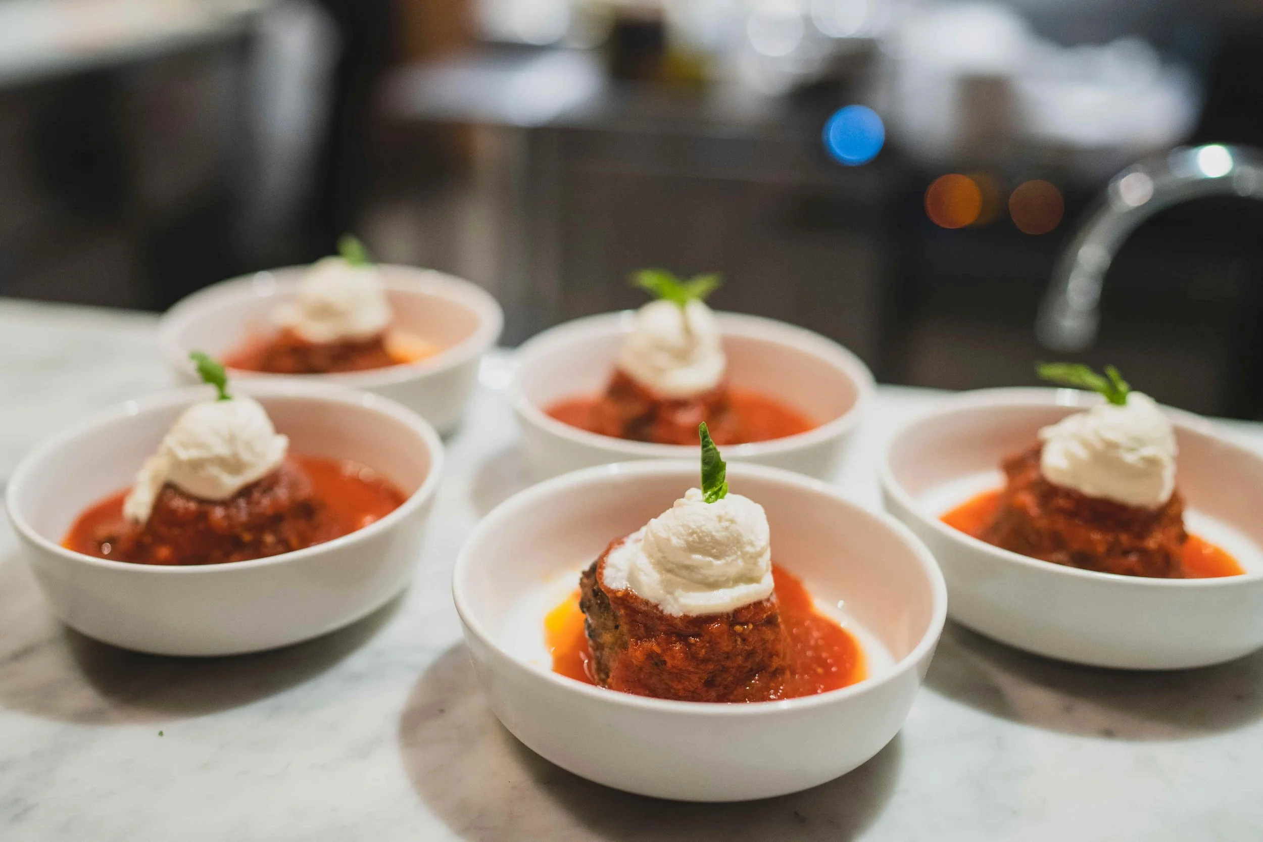 Small white bowls filled with meatloaf topped with whipped potatoes and garnished with a small sprig of basil, placed on a kitchen counter with a blurry background.