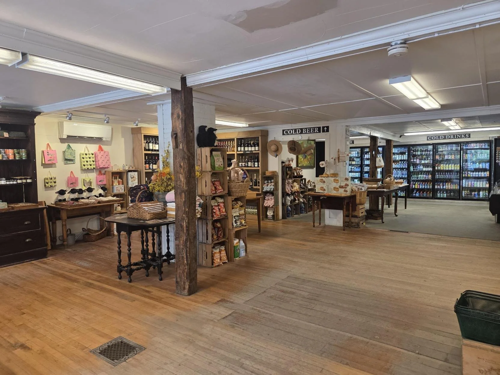 Interior of a rustic retail store with wooden floors, displayed merchandise including hats, bottles, bags, and stuffed animals, with signs indicating cold beer and cold drinks areas.